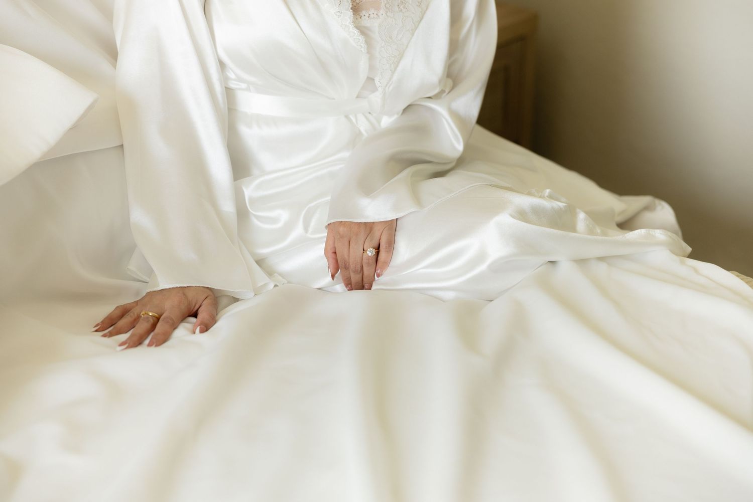 Bride in white satin wedding gown sitting on bed, elegant long sleeves and flowing skirt detail.