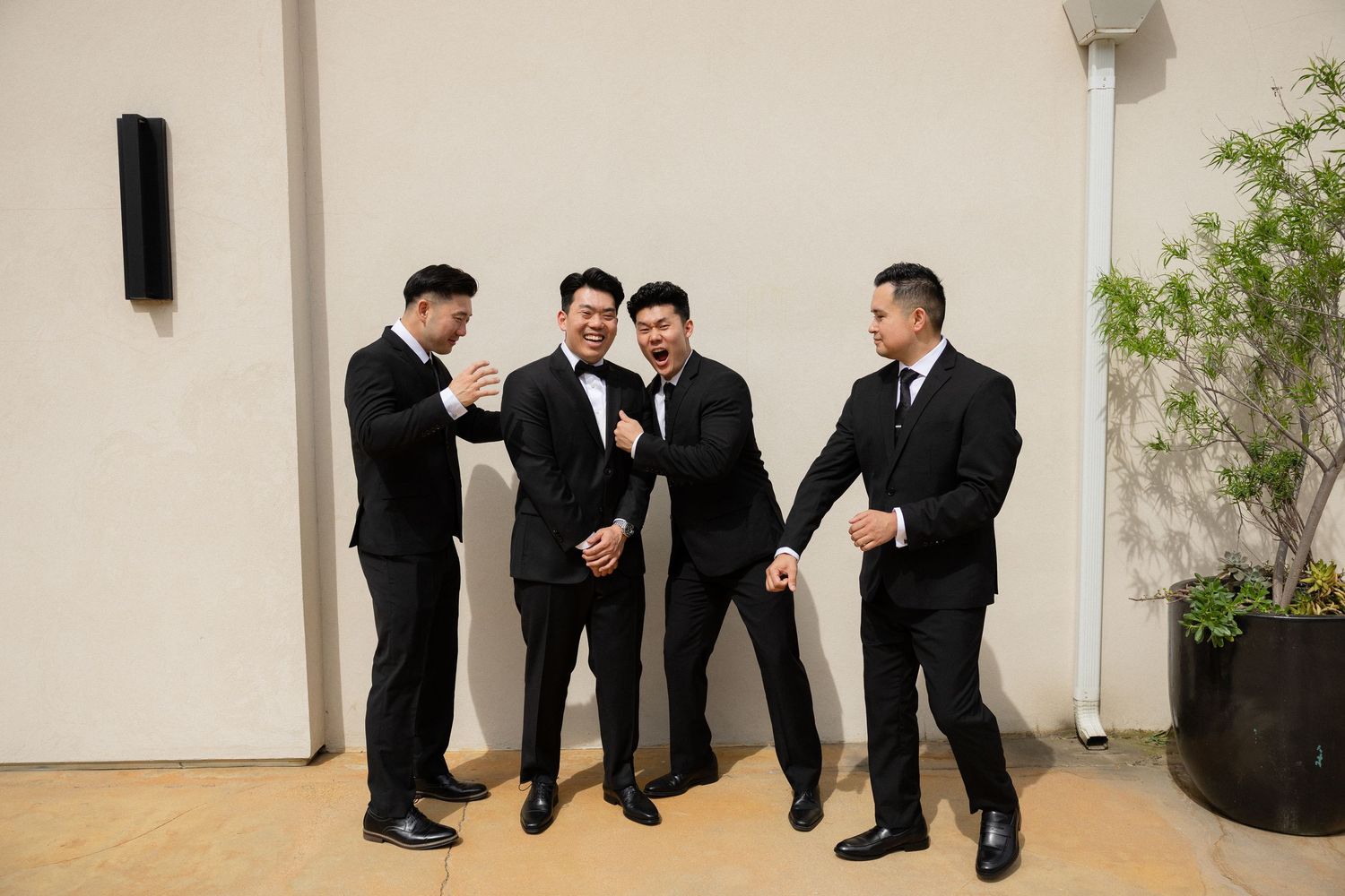 Four groomsmen laughing and celebrating together in black suits against a white wall at a wedding.