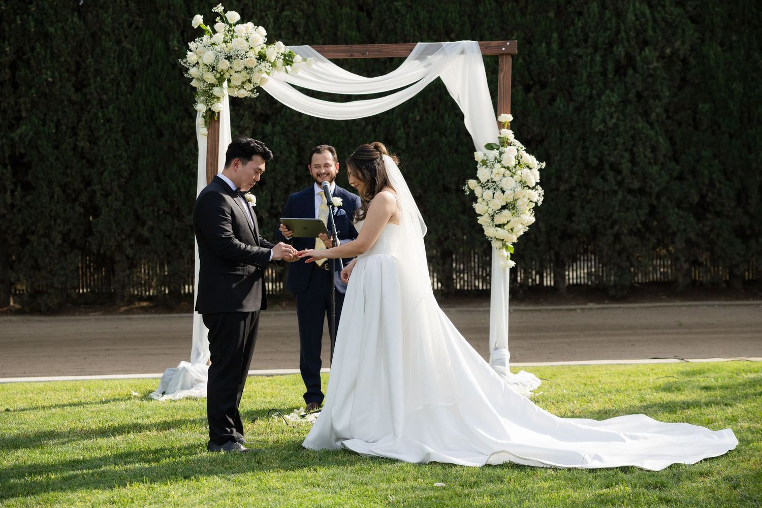 Couple exchanges vows under white draped arch adorned with white roses at outdoor ceremony.