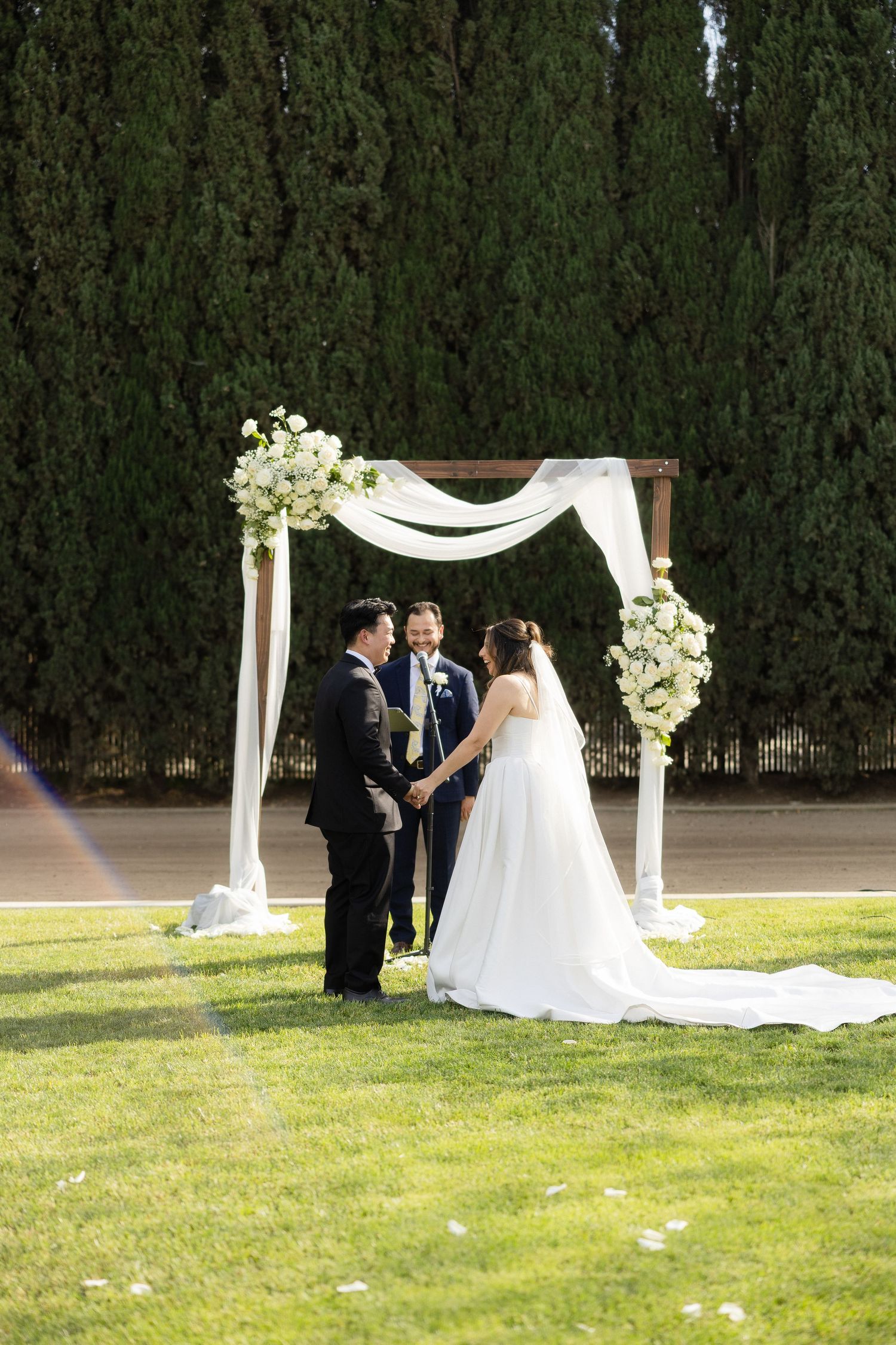 Bride and groom exchanging vows under floral wedding arch with white draping outdoors.