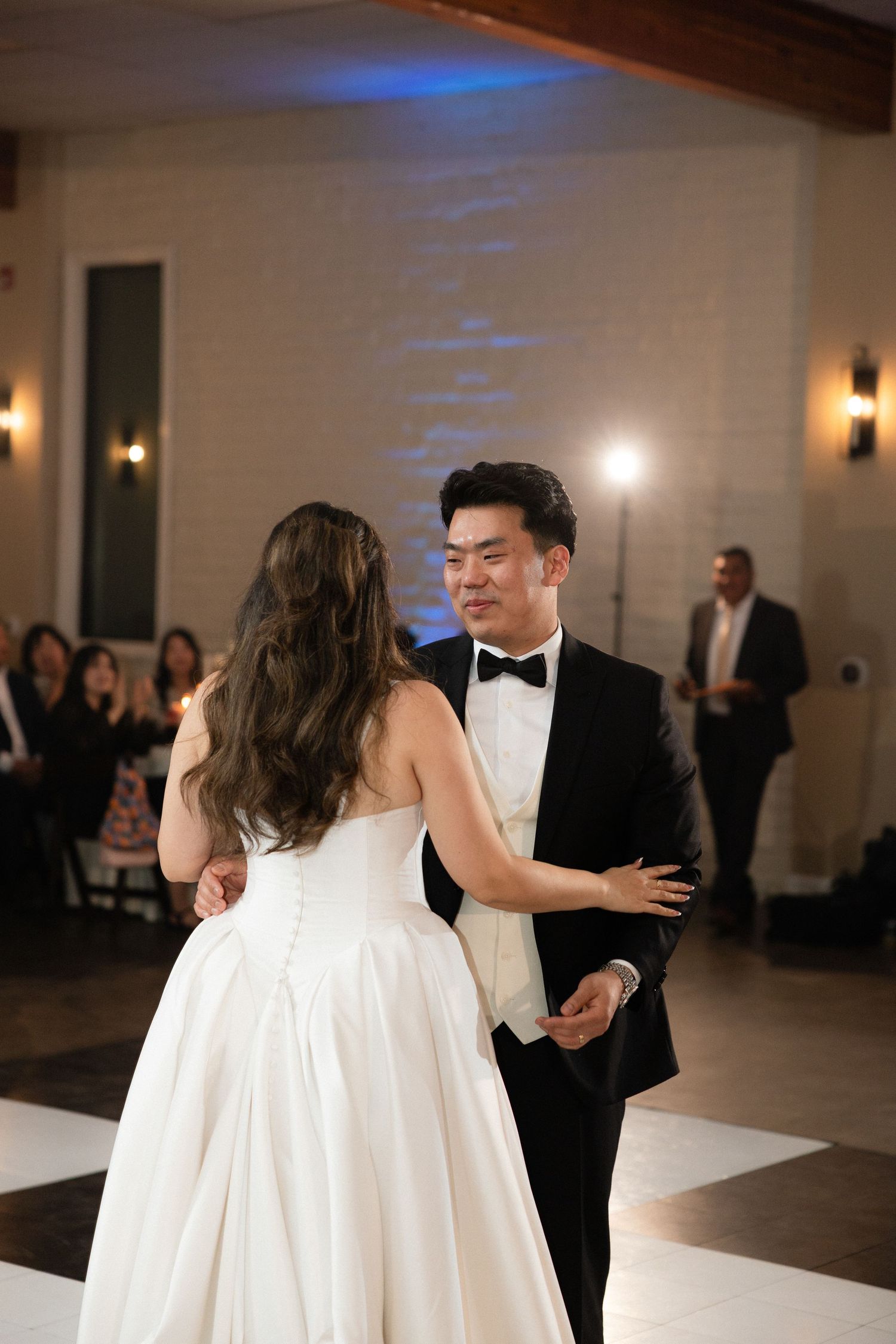 Groom in tuxedo shares first dance with bride in white gown at wedding reception.