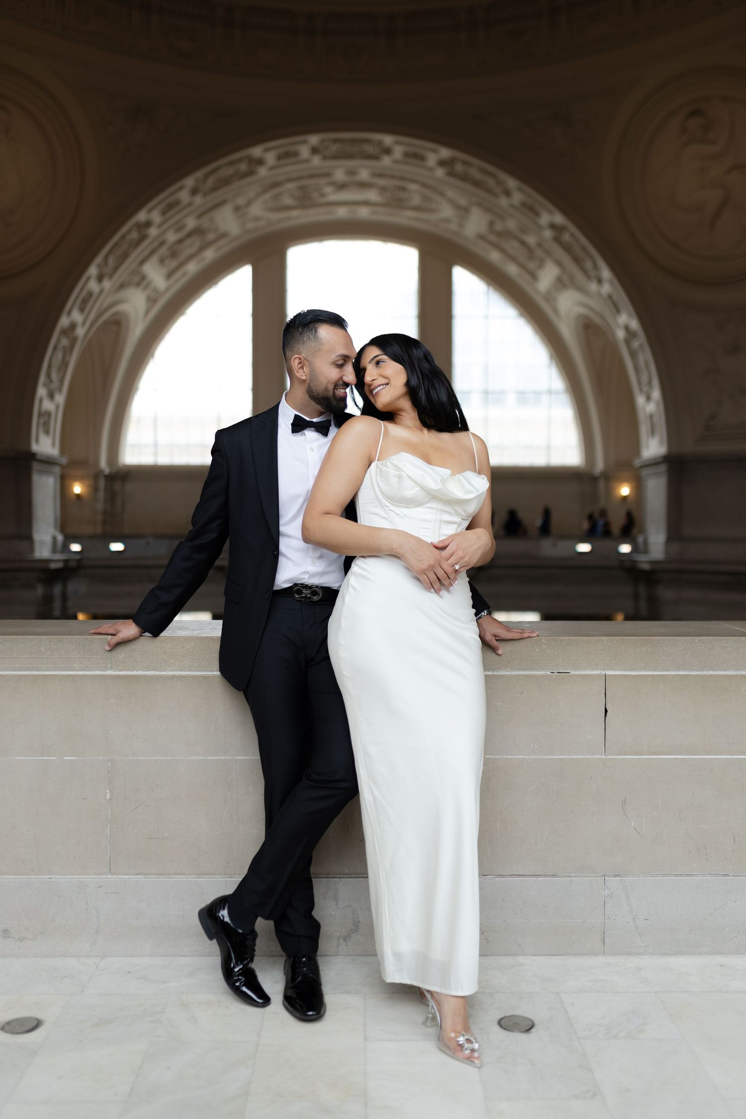 Elegant couple in formal attire pose together under an ornate arched doorway in a grand building interior.