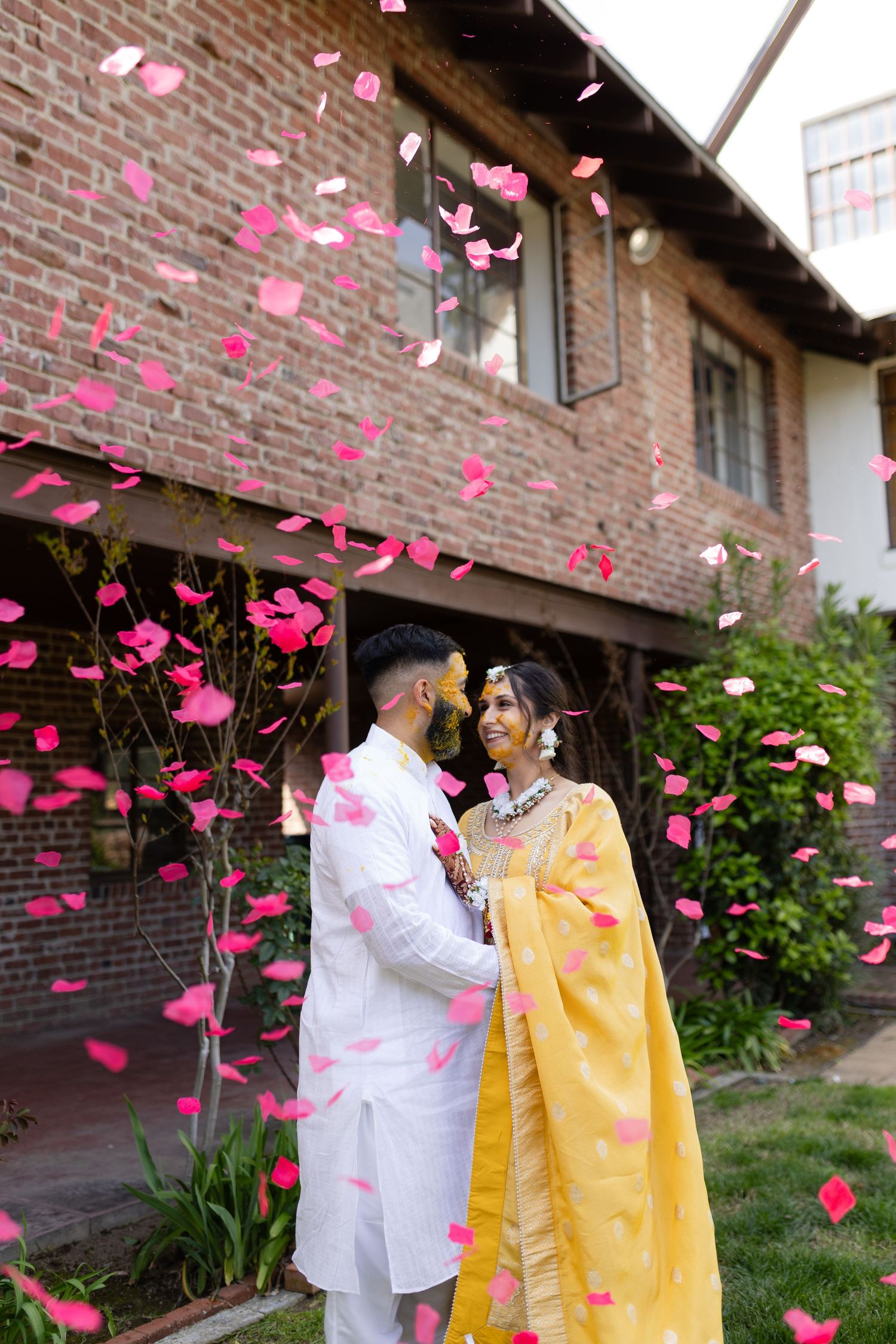 Couple in yellow and white attire showered with pink rose petals against a brick building.