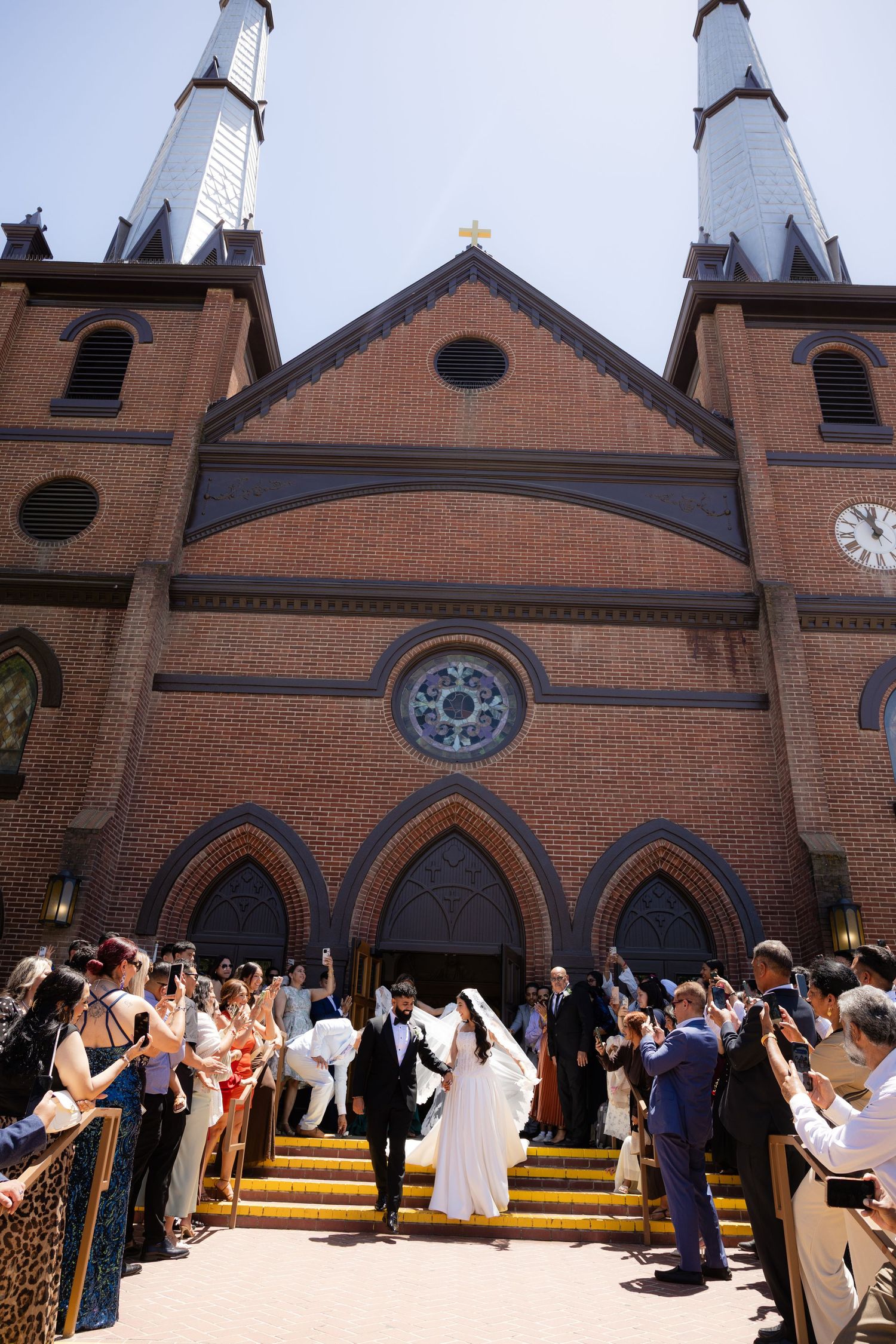 Bride and groom exit brick Gothic church flanked by cheering guests taking photos on sunny wedding day.