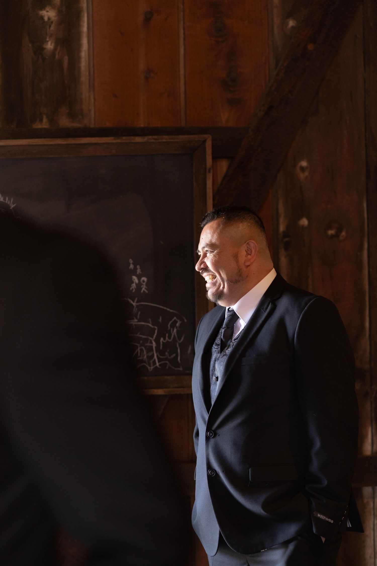 Man in dark suit stands in rustic wooden interior with dramatic lighting.