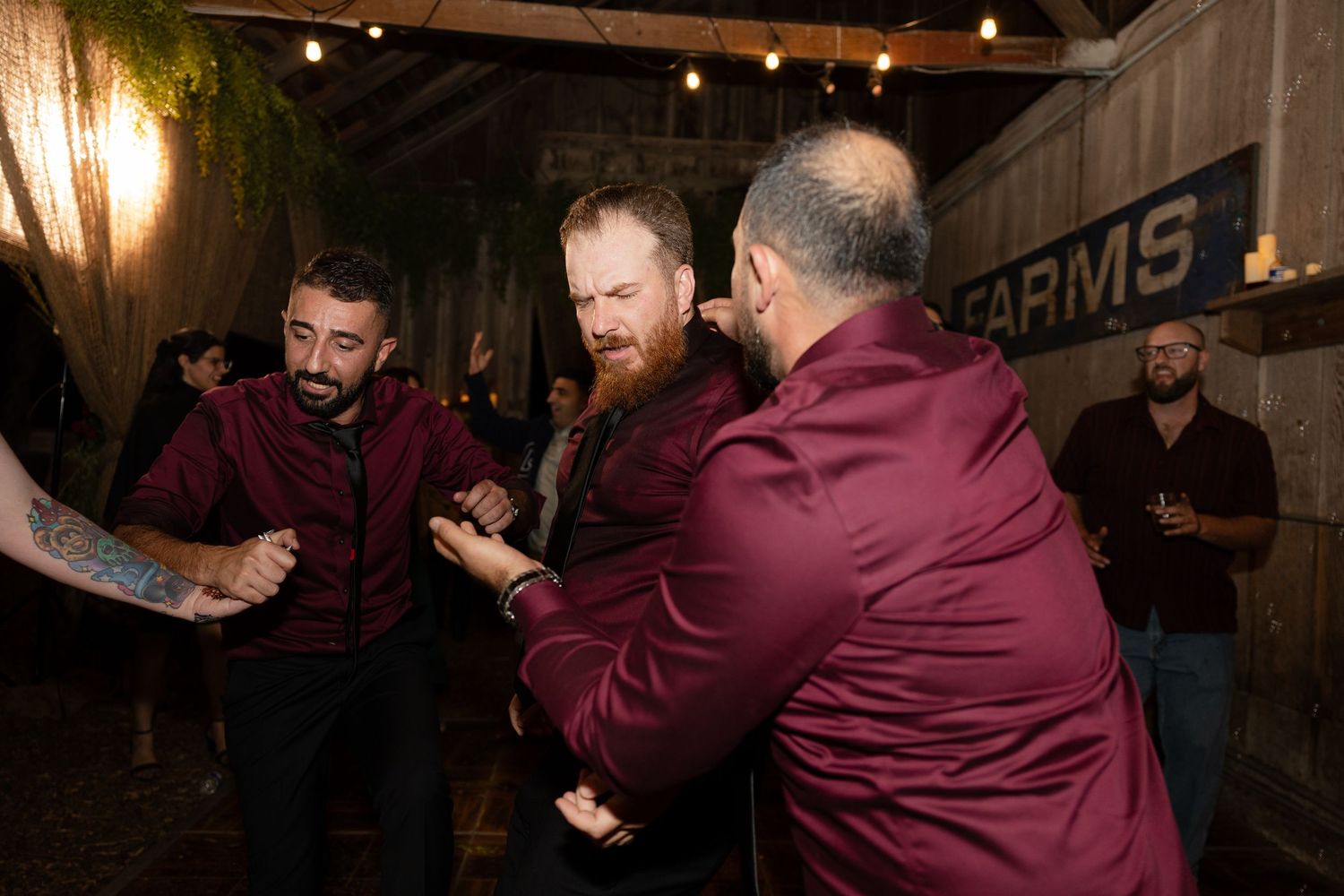 Three guests in burgundy shirts dancing together at rustic barn wedding reception.