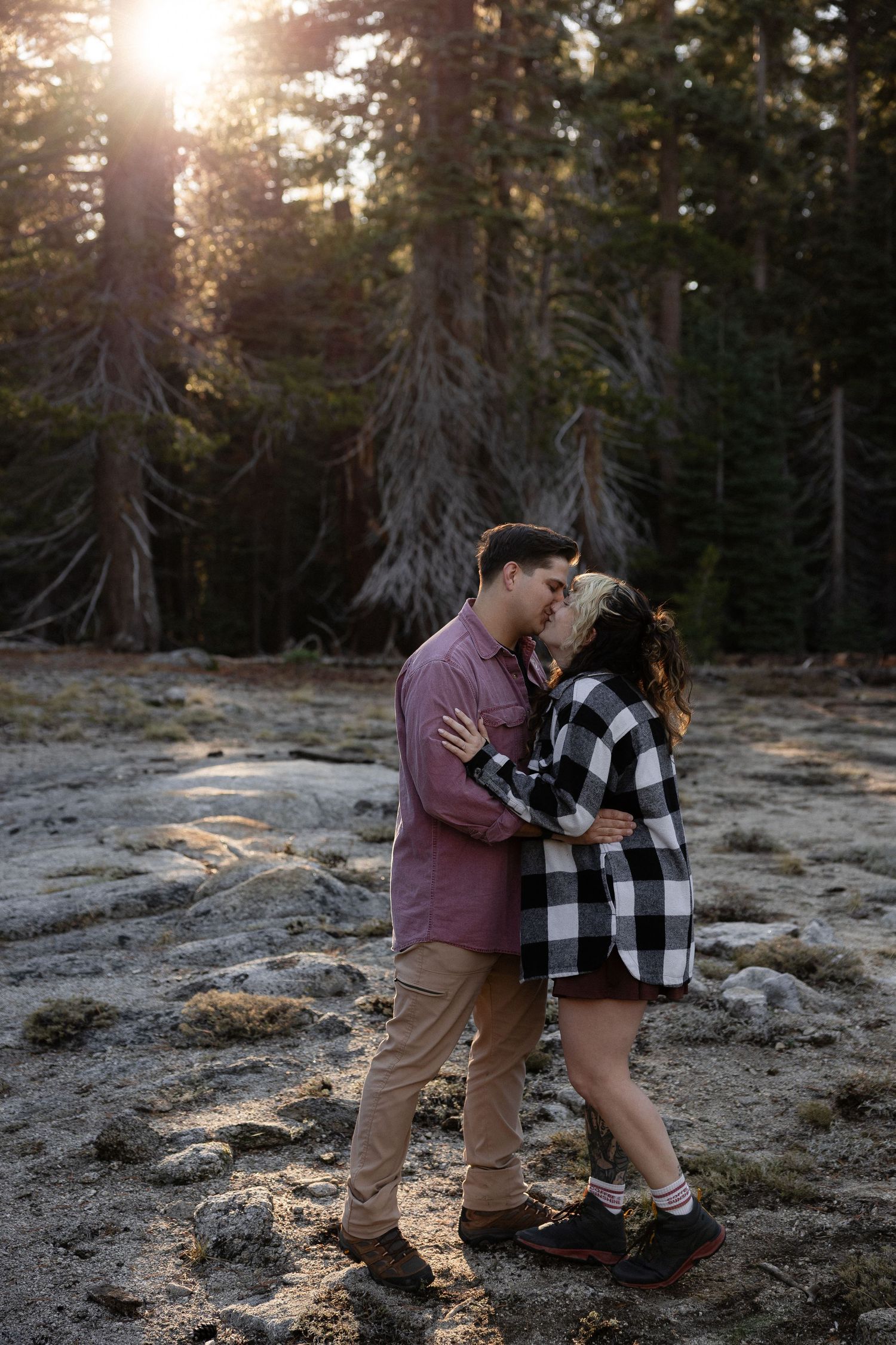 A couple sharing an intimate moment on rocky terrain with sunlit forest behind them.