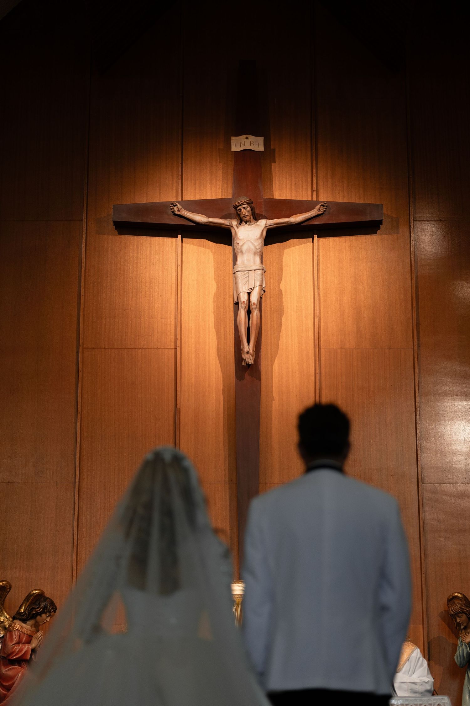 A large crucifix illuminated against wooden paneling in a church as a couple stands before it during a ceremony.