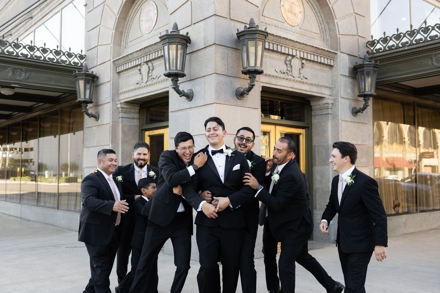 Groomsmen in formal attire posing playfully at outdoor wedding venue.