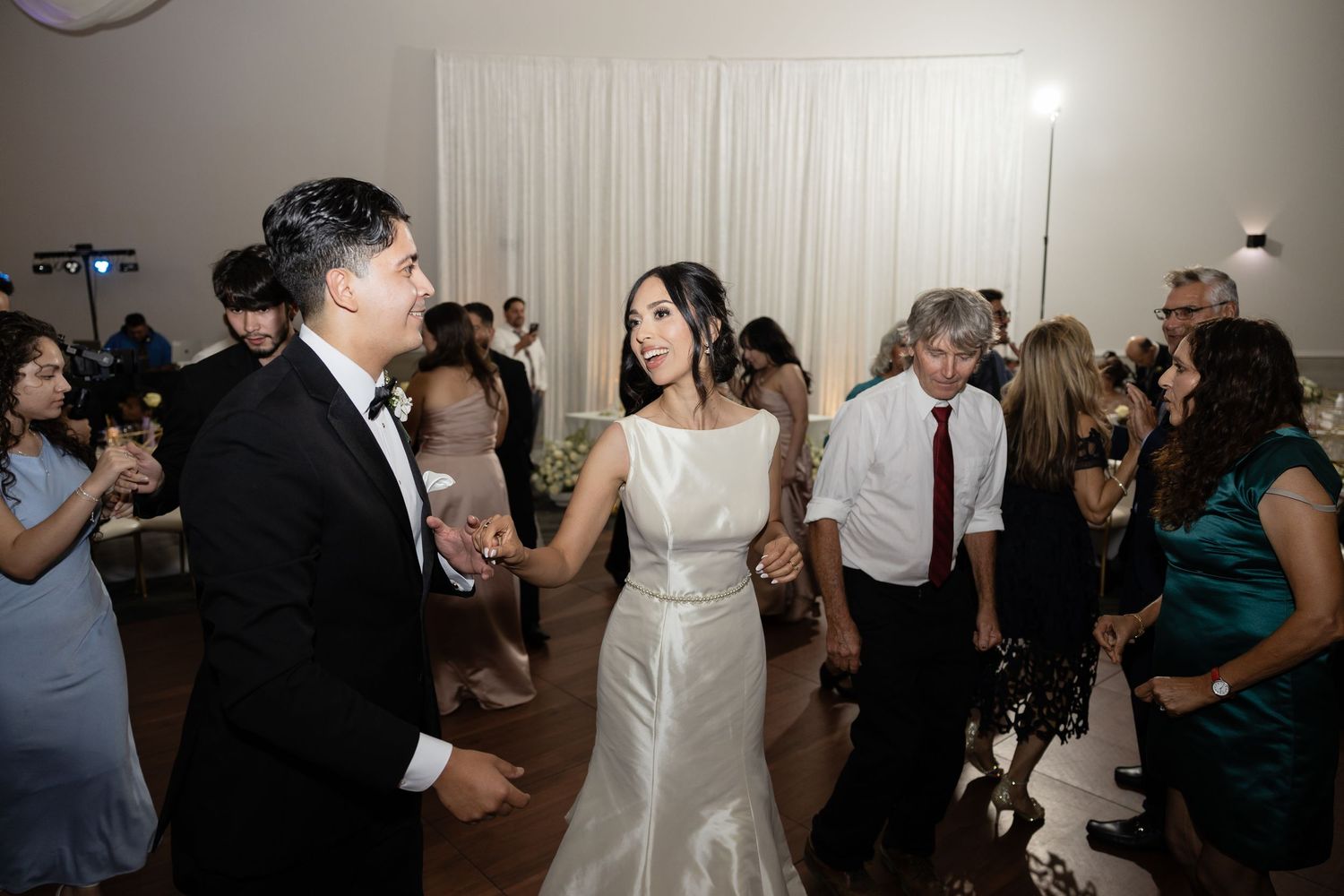 Newlyweds and wedding guests dance together on a dimly lit dance floor at an elegant reception celebration.