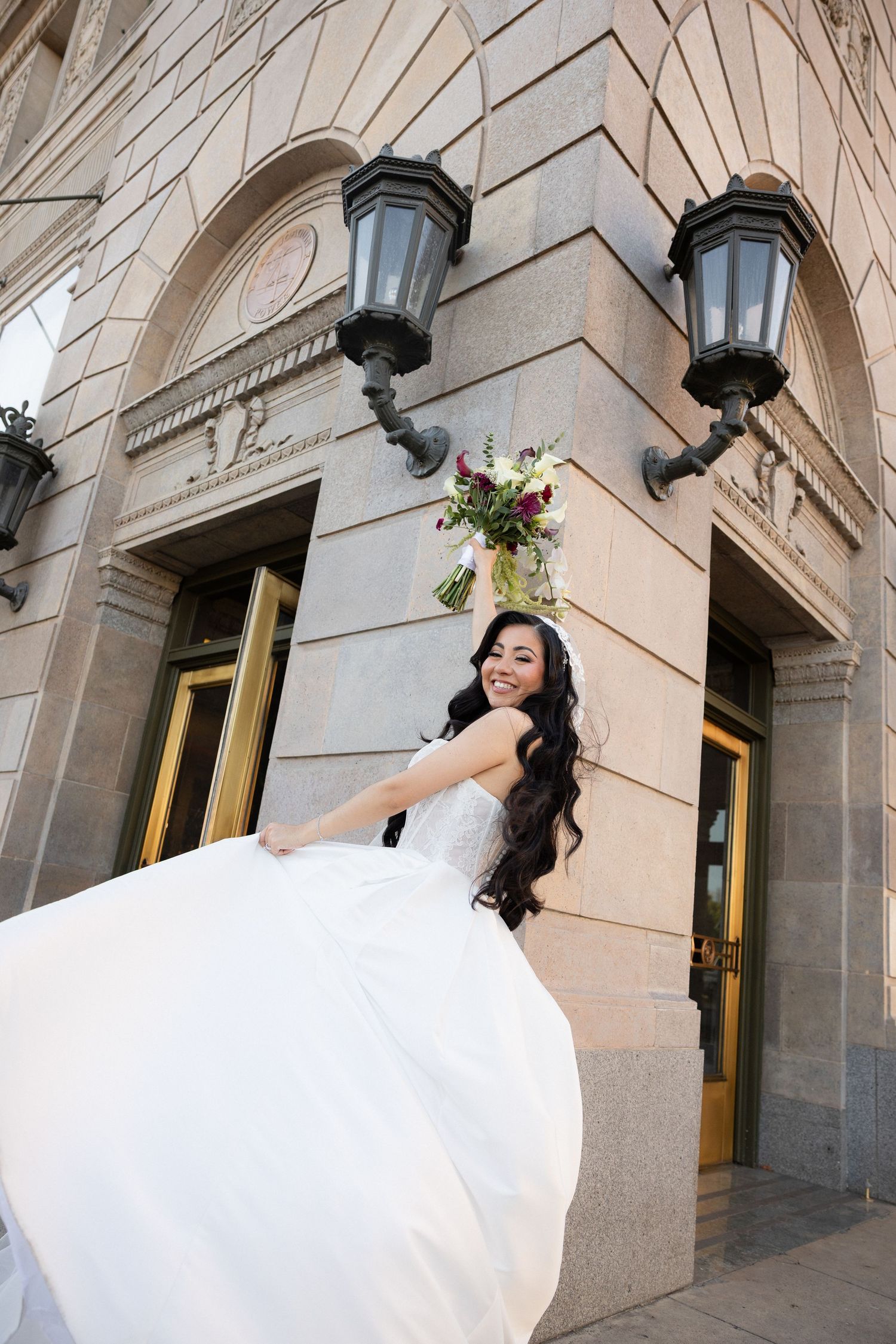 Smiling bride in a flowing white ball gown twirling outside an elegant stone building with black lanterns and a colorful bouquet.