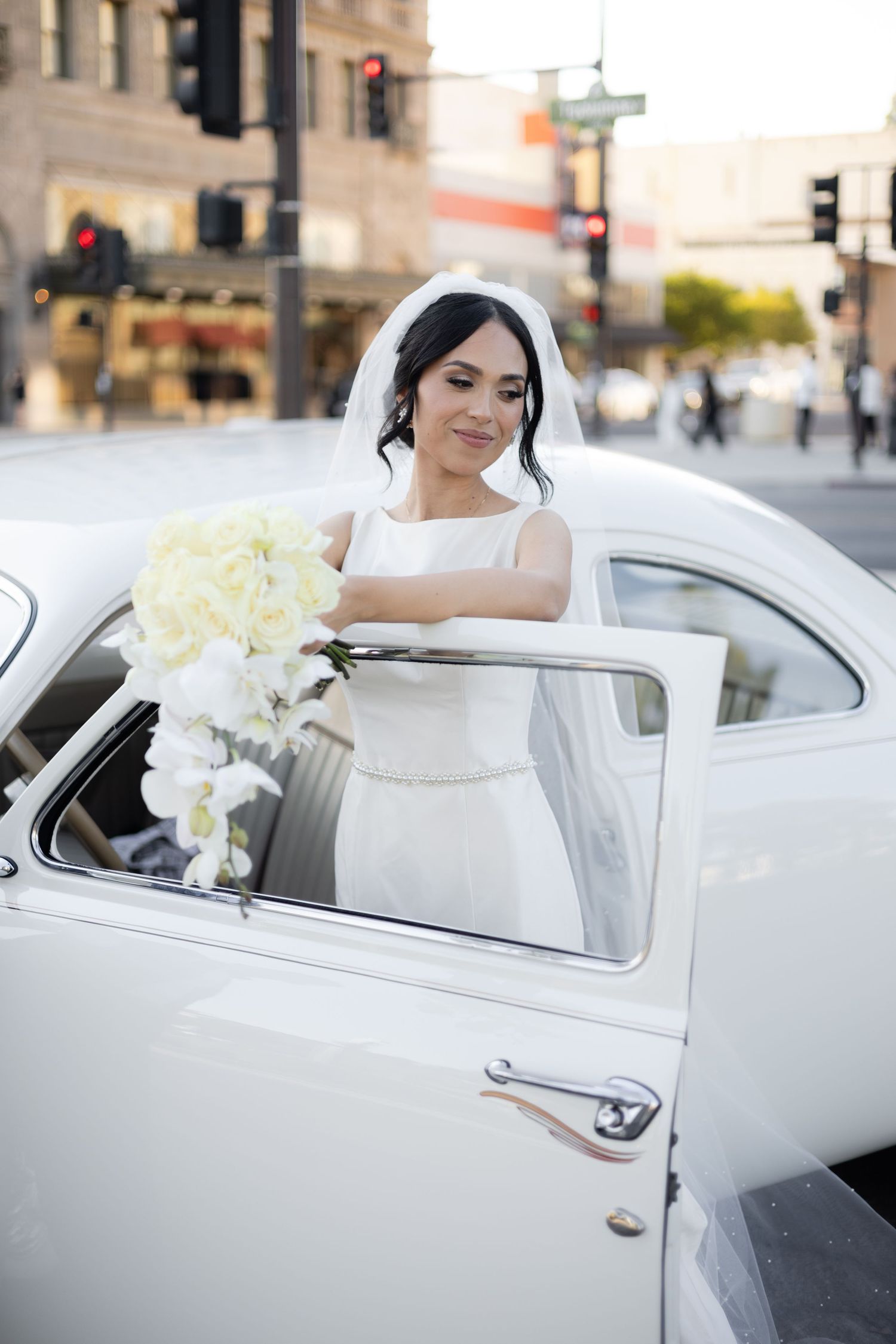 Bride in white dress sits elegantly in vintage car decorated with white roses.