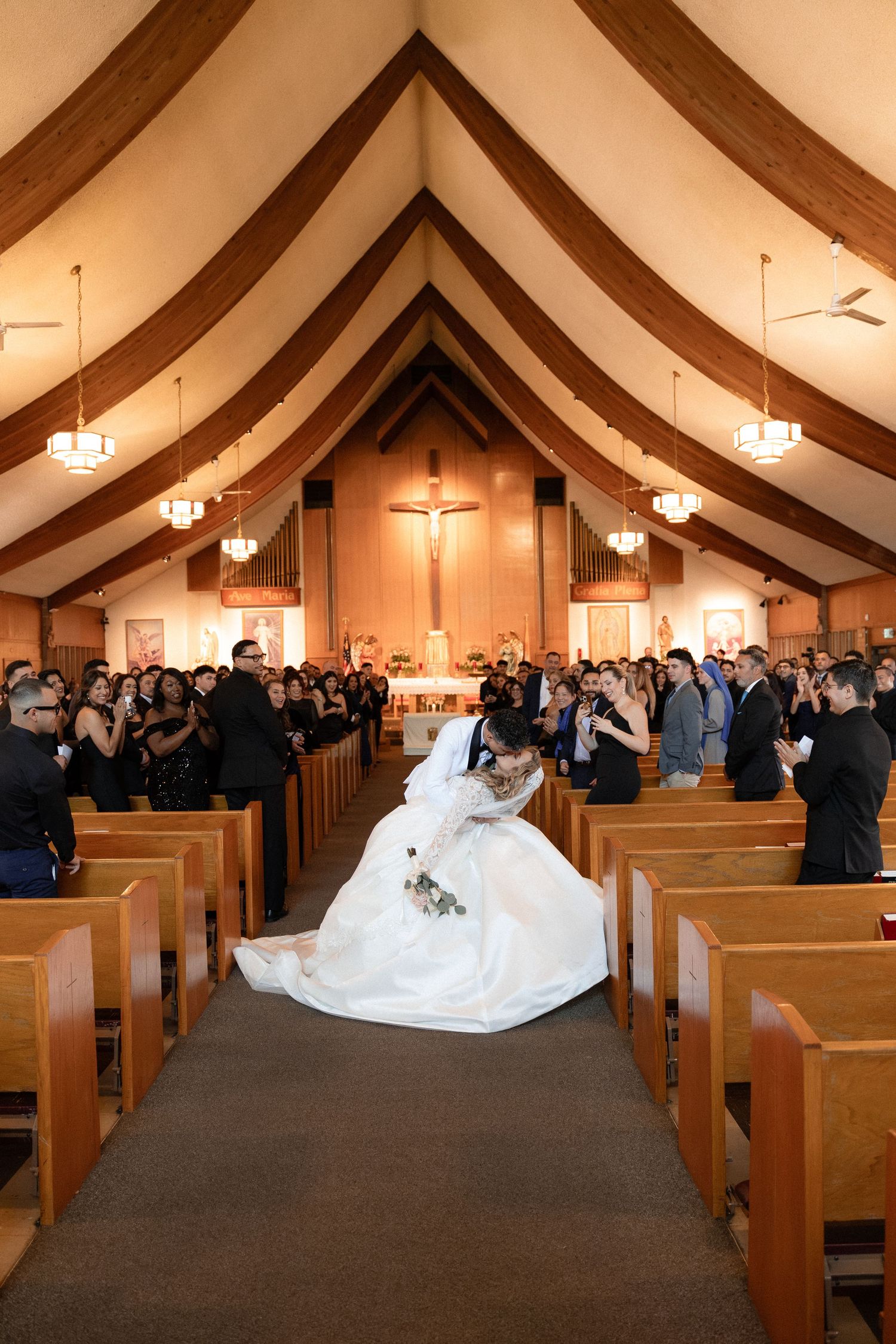Wedding ceremony in church with wooden beams and hanging lights as guests fill the pews.