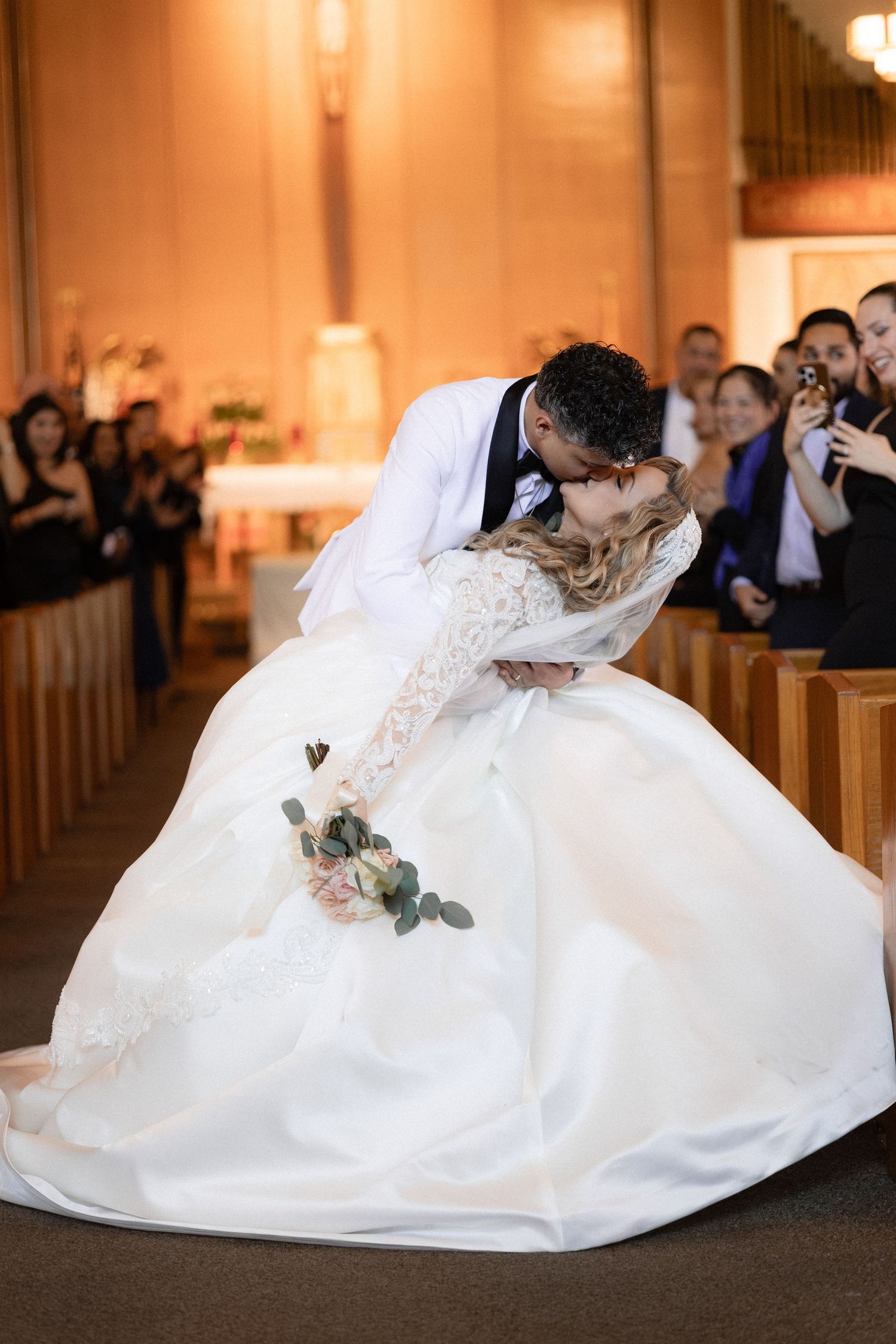 Couple shares romantic dip kiss in church with guests watching from wooden pews.