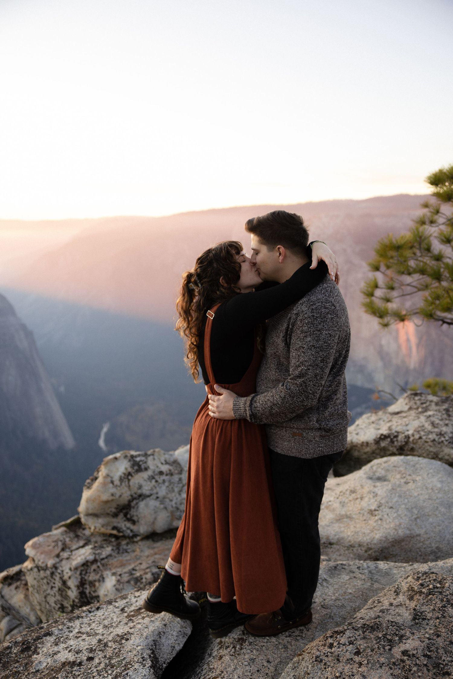 A couple in rustic attire shares a tender moment on a mountain overlook during golden hour.