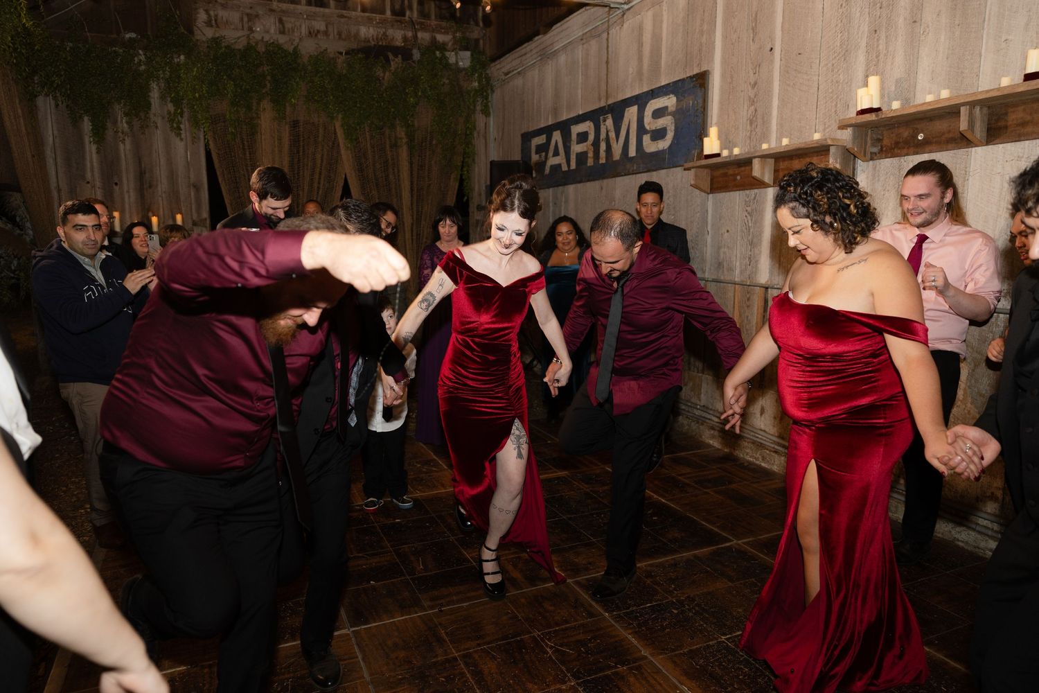 Wedding party in burgundy attire dances energetically in rustic barn setting.