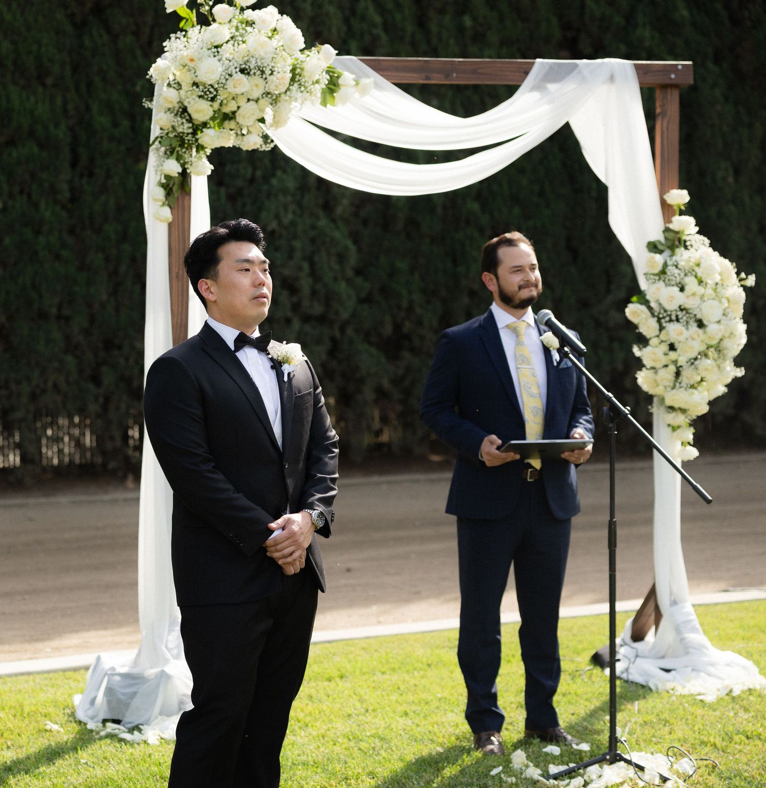 Groom in black tuxedo stands at white floral wedding arch while officiant reads ceremony script.