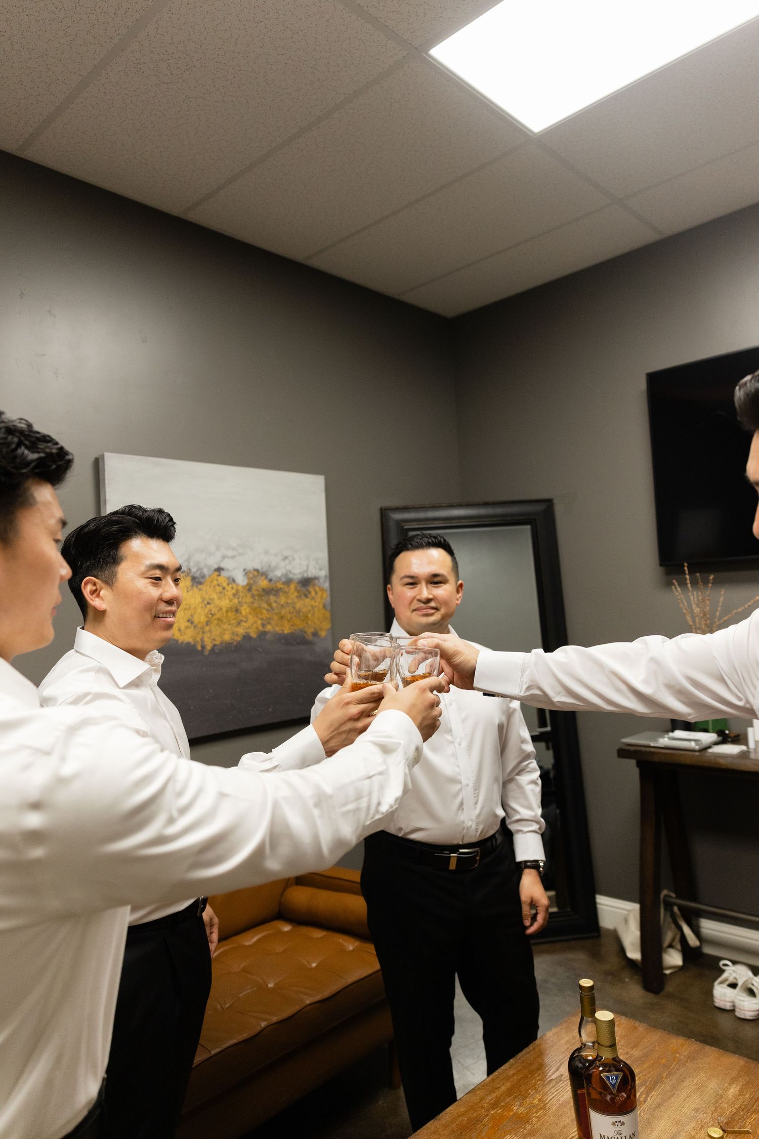 Groomsmen in white shirts toasting with glasses of whiskey in a modern gray room before a wedding.