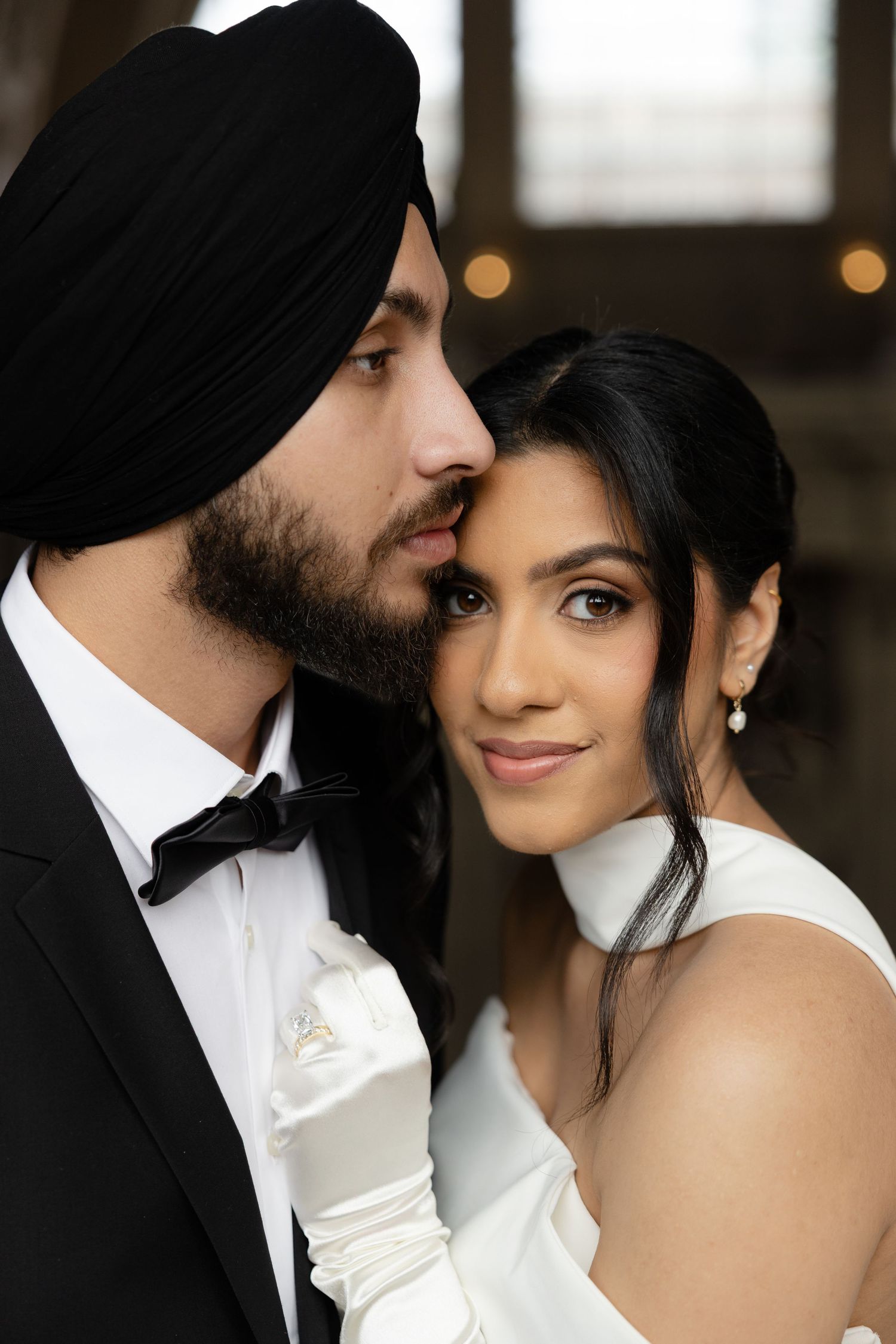 Elegant couple in formal wedding attire share an intimate moment, with the groom wearing a black turban and bow tie.