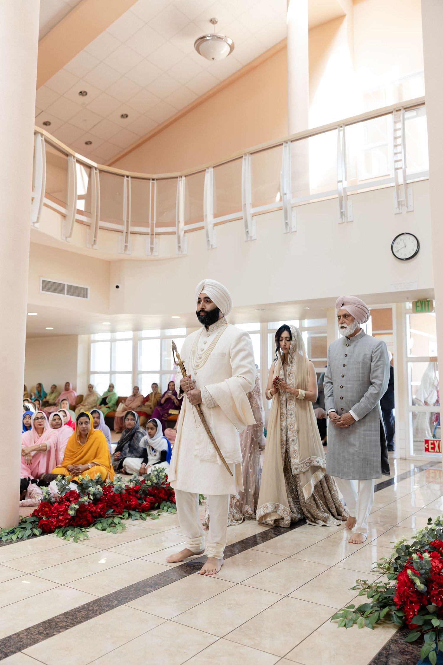 Traditional Sikh wedding ceremony taking place in a bright modern Gurdwara with red floral decorations.