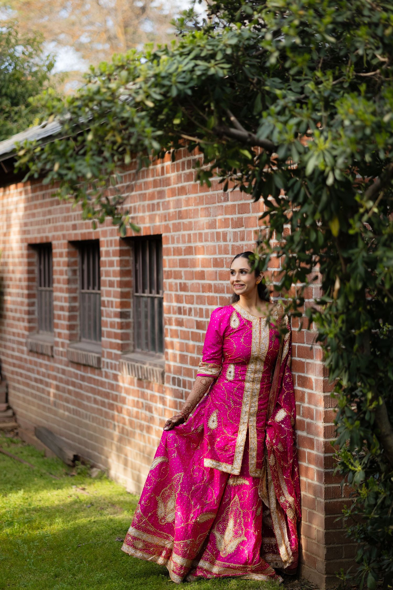 Traditional pink and gold Indian sari dress flows elegantly against rustic brick wall backdrop with green foliage.
