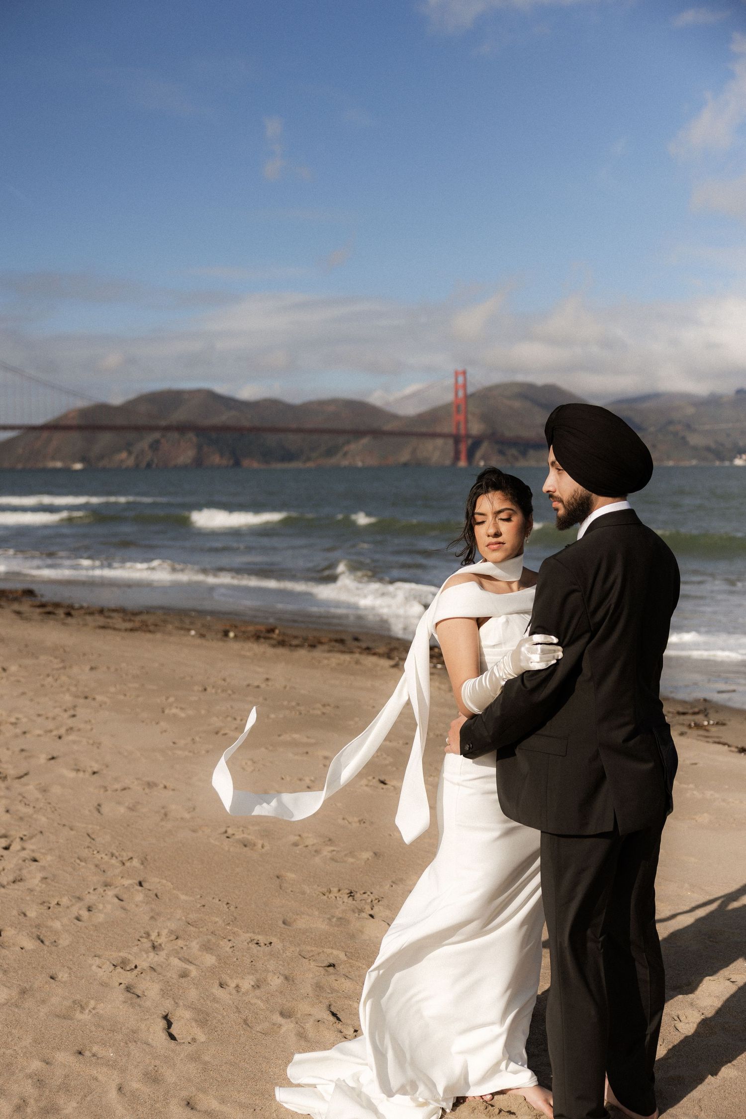 A couple dances on Baker Beach with the iconic Golden Gate Bridge visible in the background on a cloudy day.