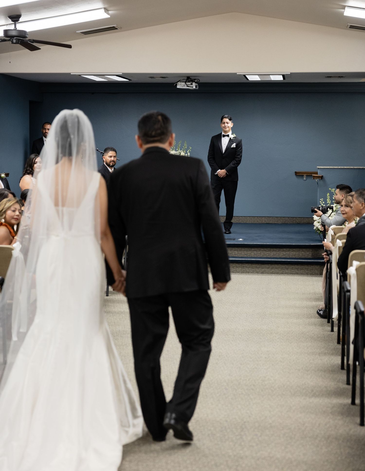 Wedding ceremony procession in a large indoor venue with rows of seating.