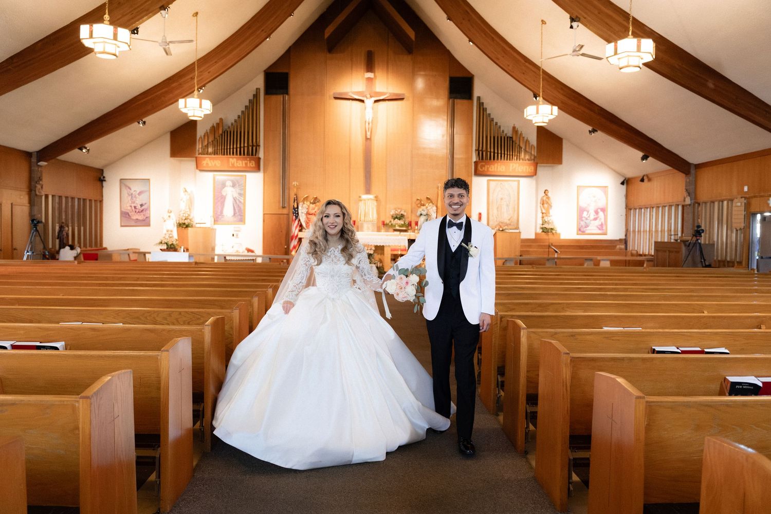 A couple stands in the center aisle of a church with wooden pews and vaulted ceiling.