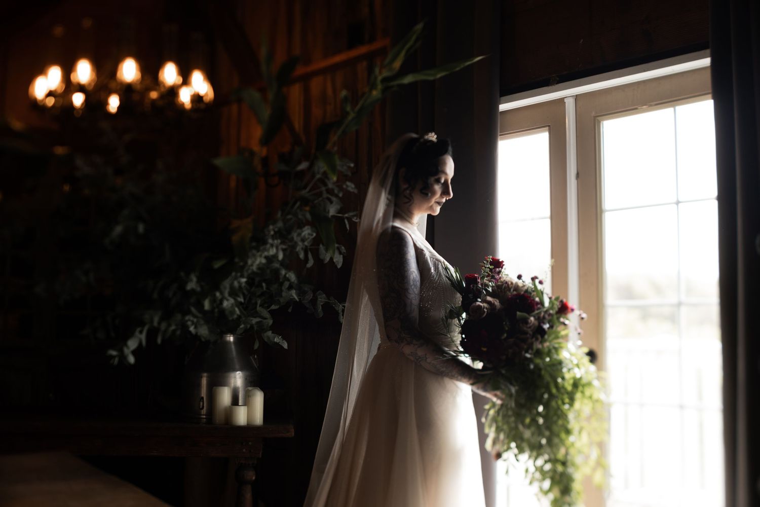 Moody wedding photography of a bride in white dress holding bouquet by sunlit window.