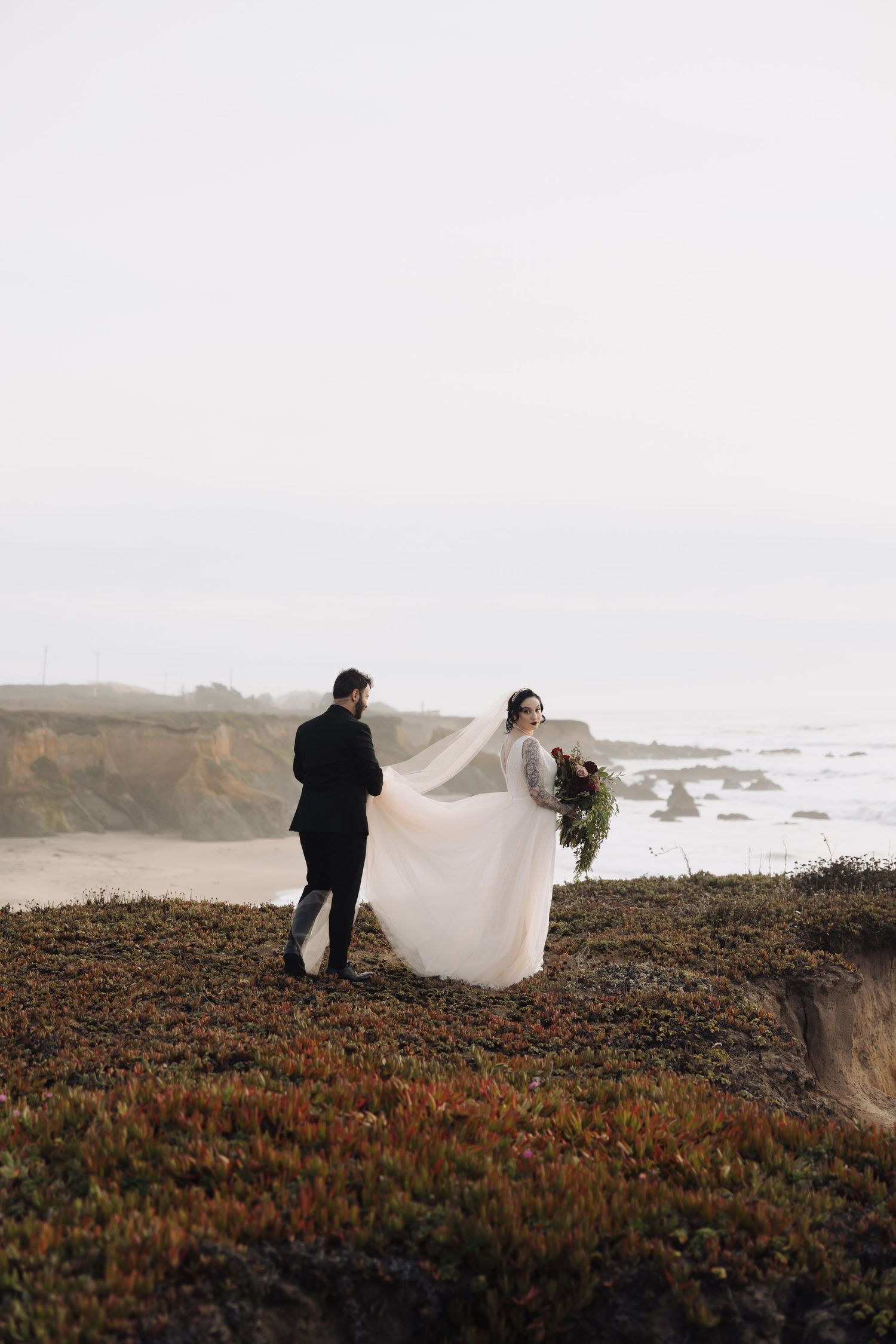 A dramatic wedding photo with flowing veil captured on rugged ocean cliffs.