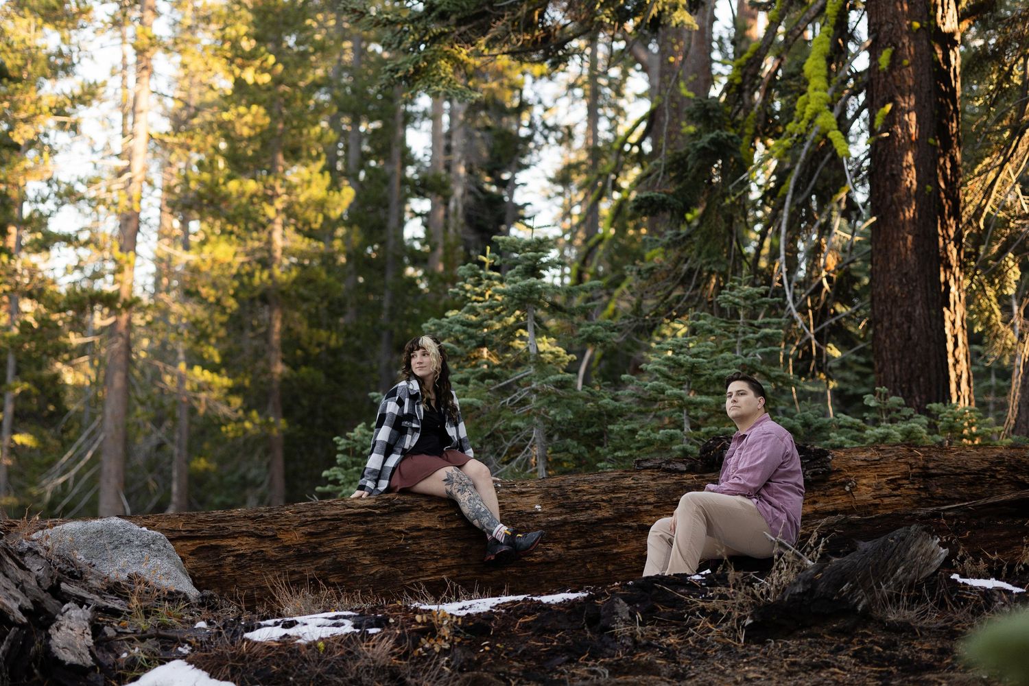 A couple explores a snowy forest trail during golden hour sunset among tall evergreen trees.