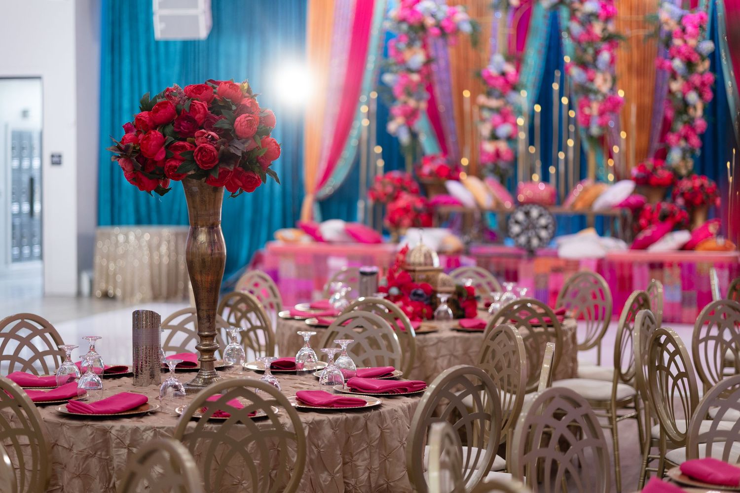 Elegant wedding reception hall with red rose centerpieces, gold chairs, and turquoise and pink drapery decor.