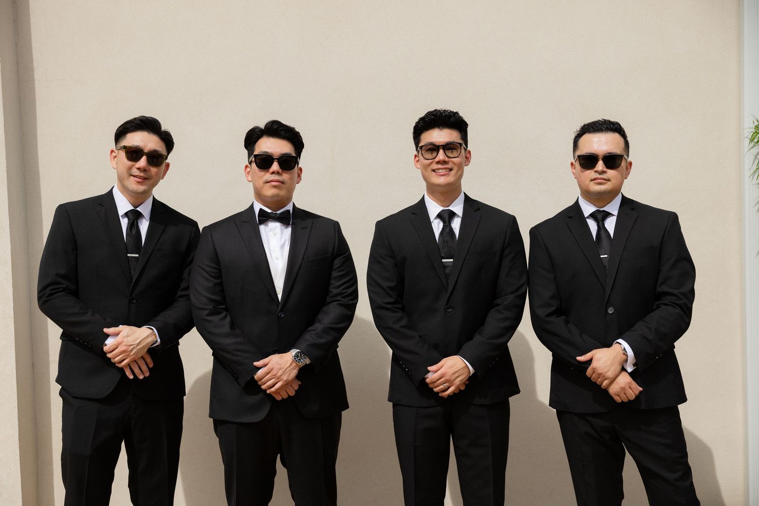 Four groomsmen wearing matching black suits and sunglasses pose together before a wedding.
