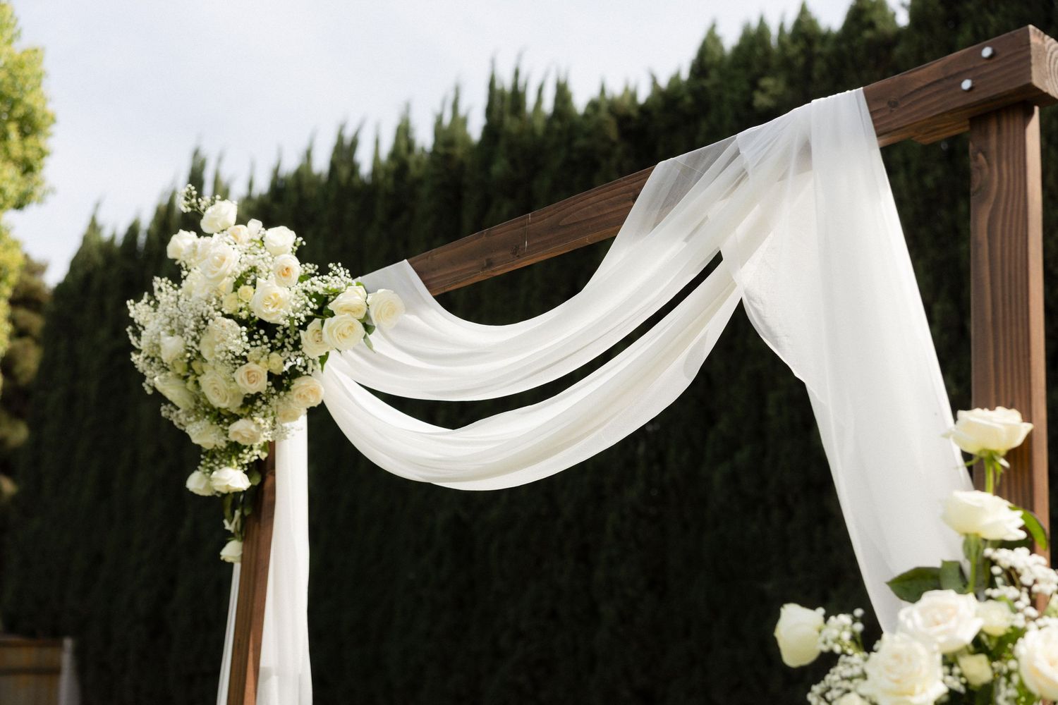 Elegant wooden wedding arch draped with white chiffon fabric and adorned with white roses and greenery outdoors.