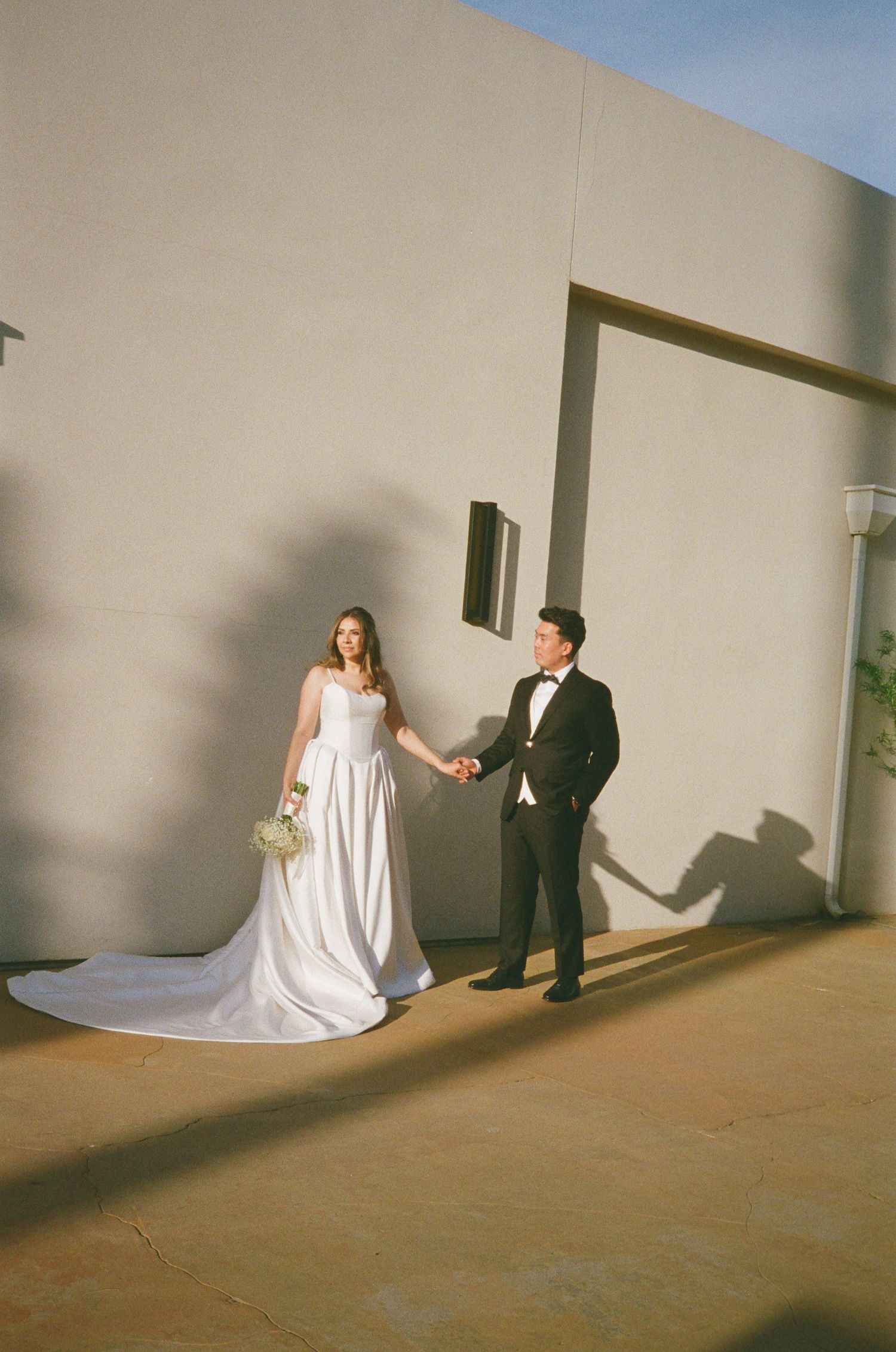 Bride in white gown and groom in tuxedo hold hands outside a sunlit building, casting shadows on the wall.