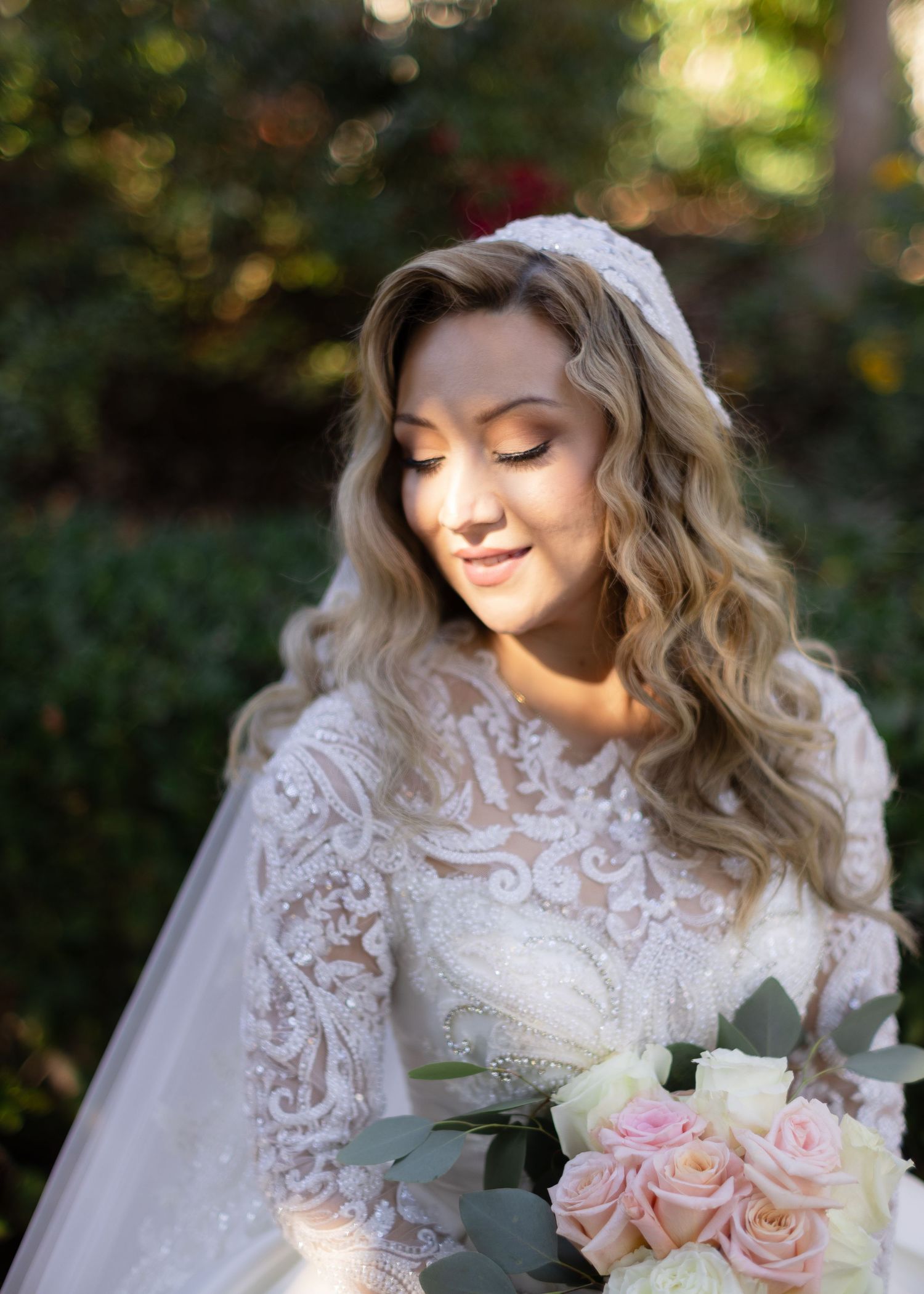 A bride in white lace smiles softly while holding a bouquet of blush roses against a natural outdoor background.