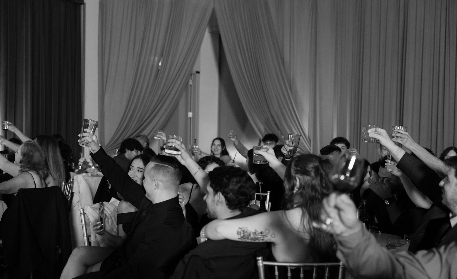 Black and white photo of wedding reception crowd with raised hands during celebration.