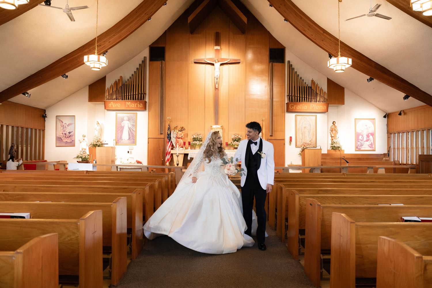 A couple walks down the aisle of a Catholic church with wooden pews and a crucifix on display.