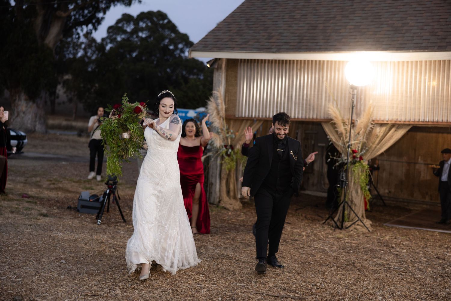 Outdoor wedding celebration at dusk with barn and string lights creating rustic ambiance.