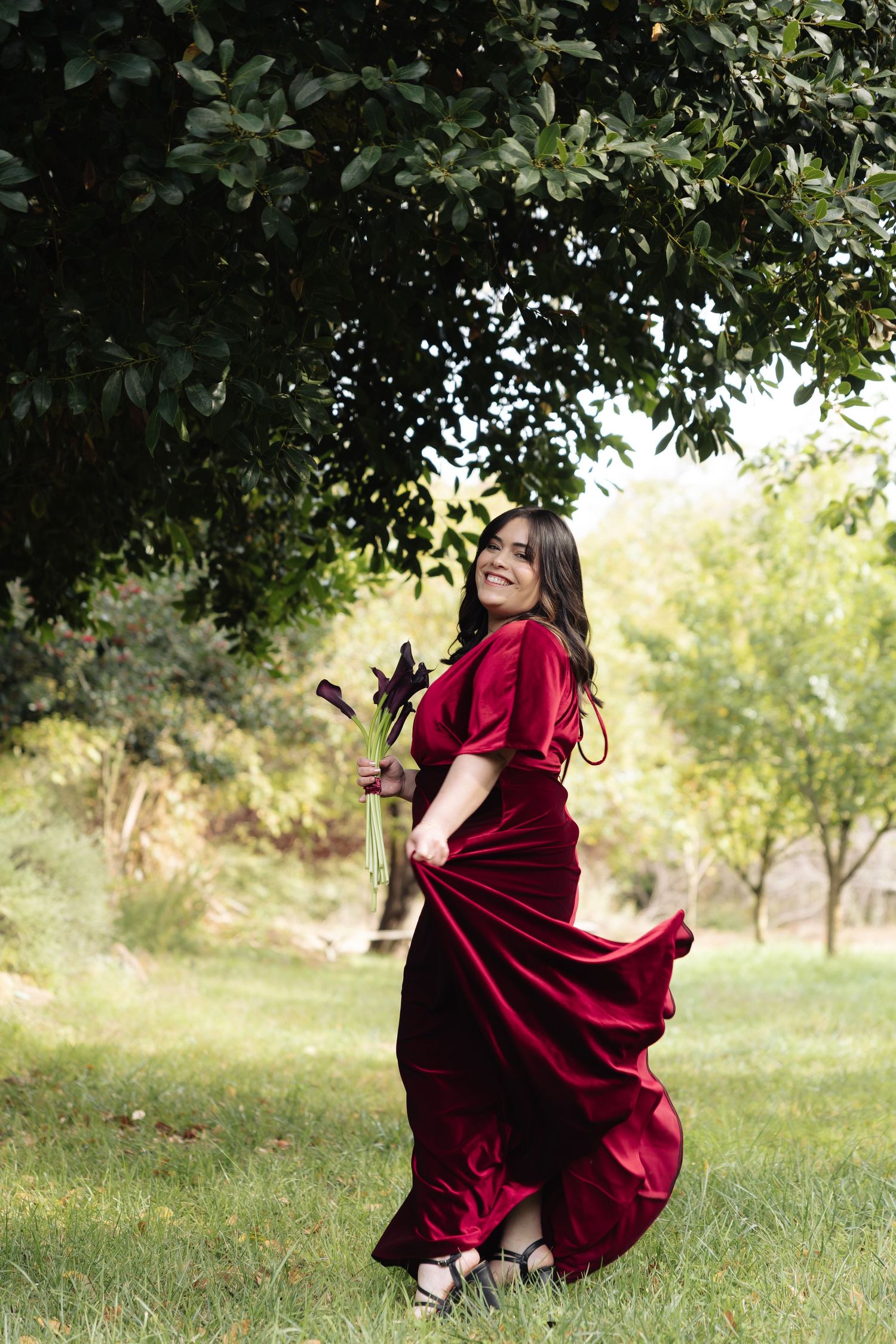 Person in flowing red velvet dress twirls gracefully in a sunlit garden setting.