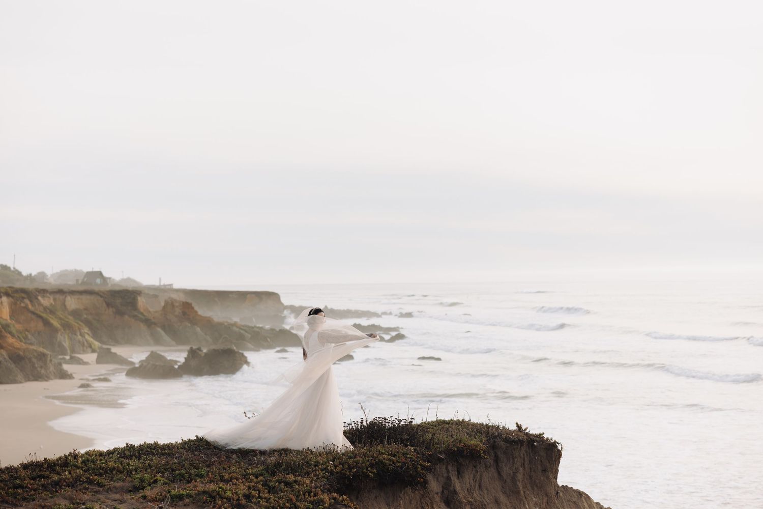 A dramatic coastal wedding photo showing a white dress flowing in the wind on rugged ocean cliffs.