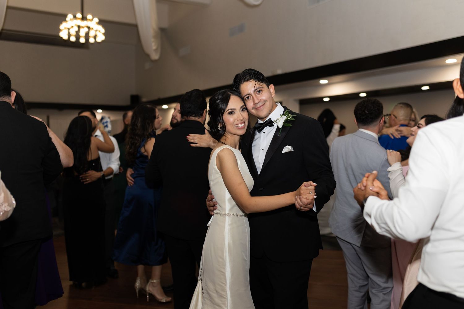 Wedding guests dance and celebrate on a dark dance floor under chandelier lighting.