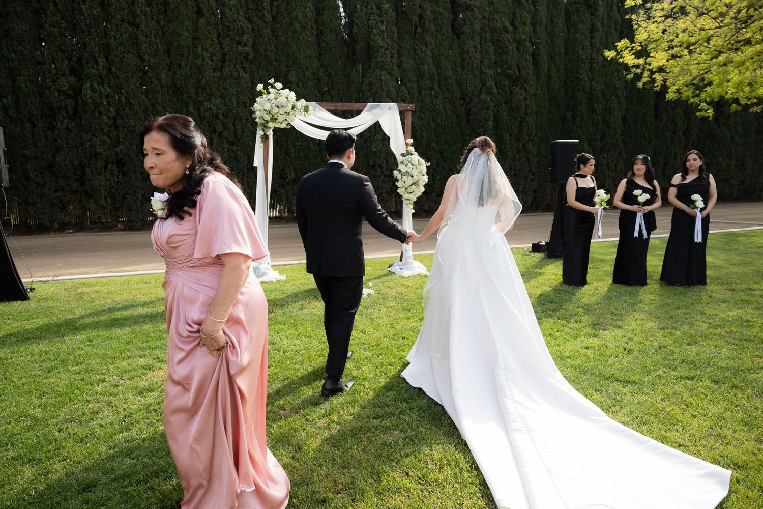 Bride and groom walk toward floral arch as bridesmaids in black dresses look on.