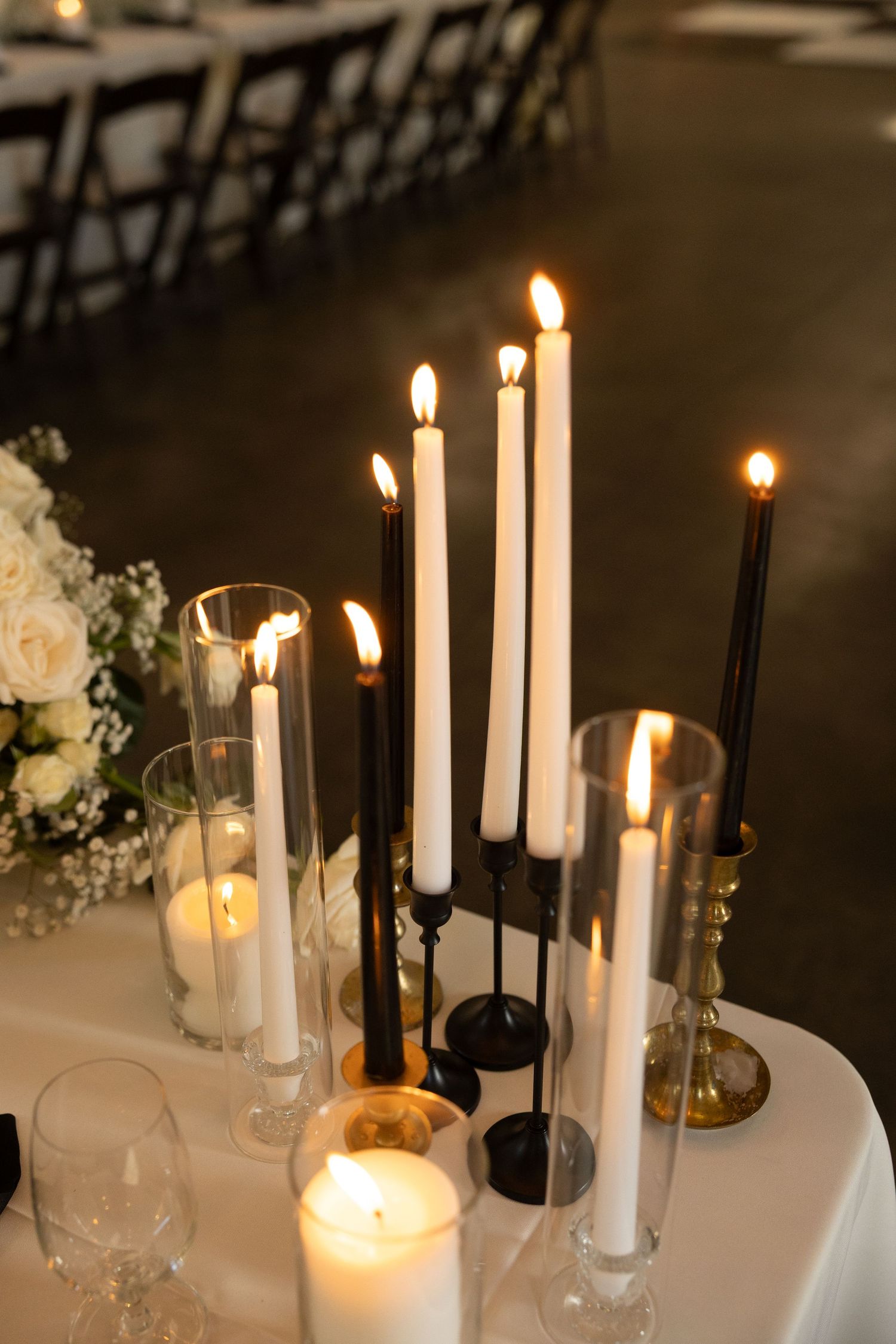 Elegant wedding reception table with lit black and white taper candles in mixed candleholders beside white floral arrangements.