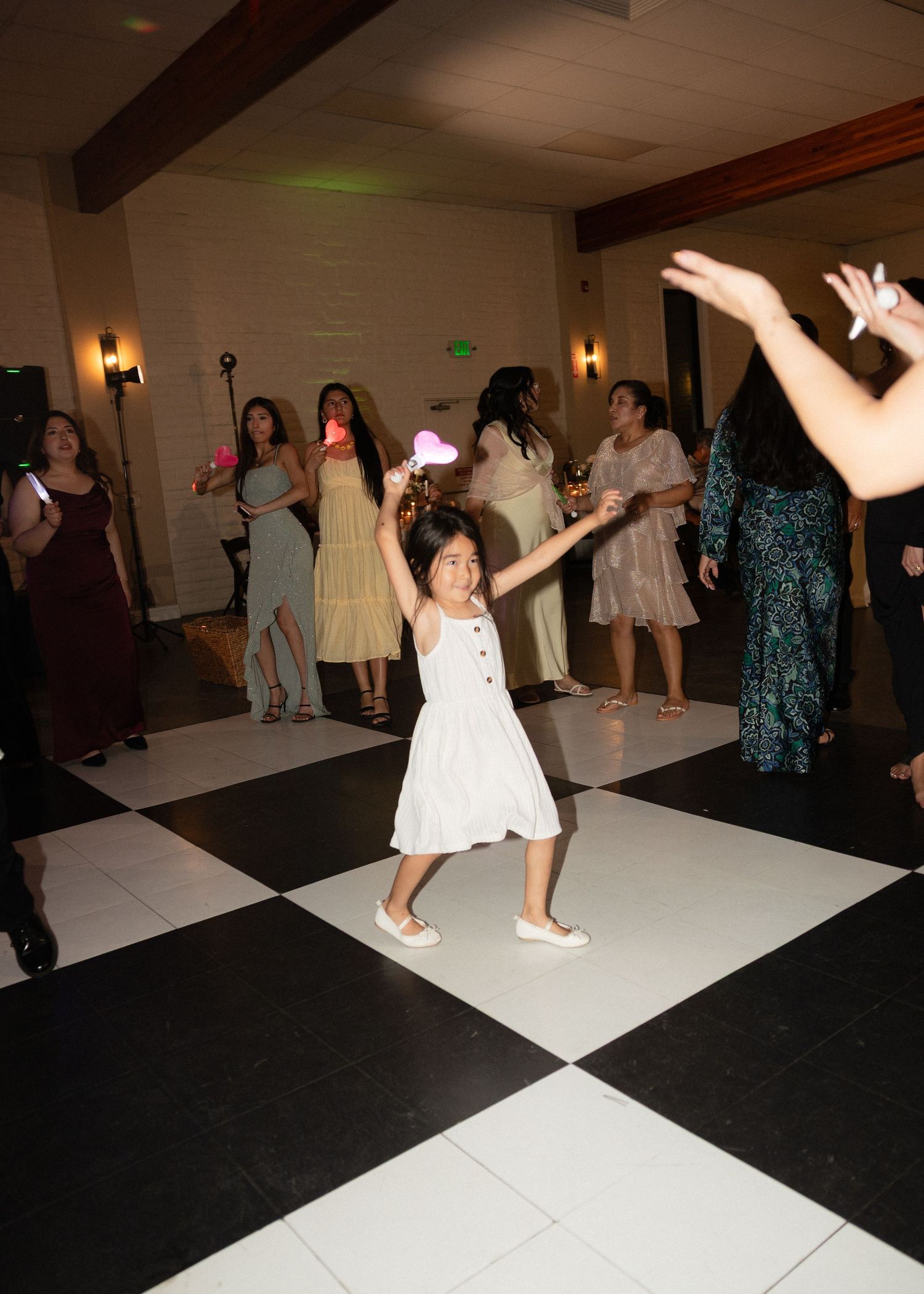 Young girl in white dress dances joyfully at wedding reception on checkered dance floor.