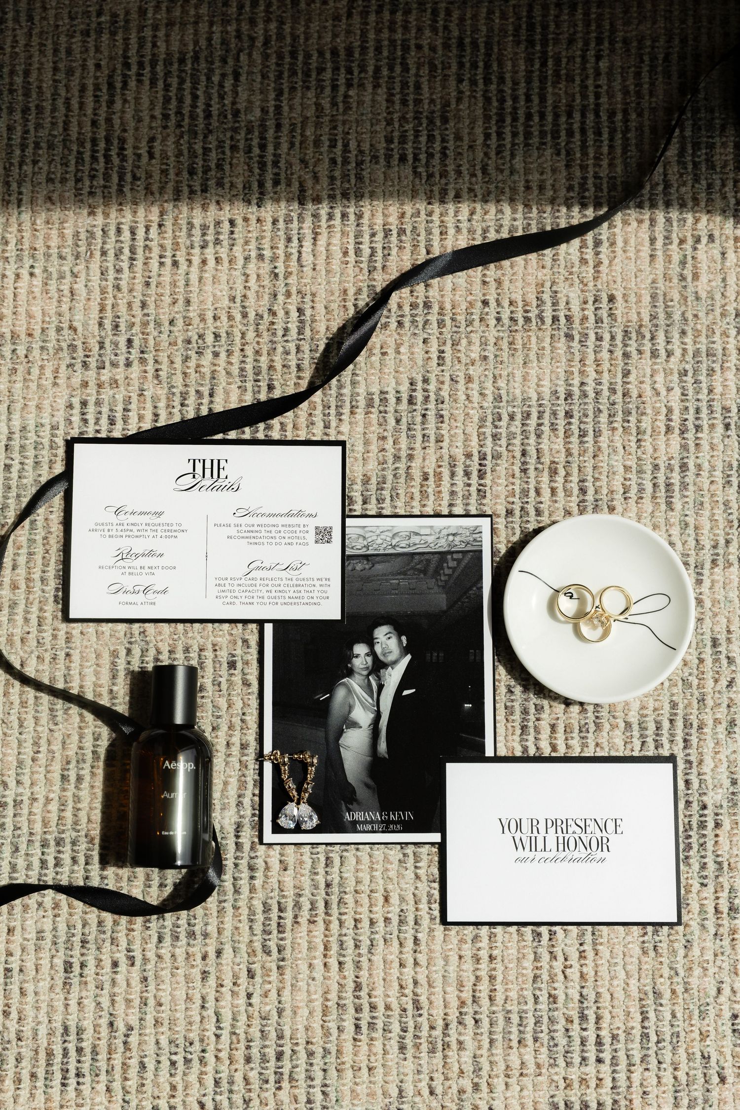 Elegant wedding flat lay with black ribbon, perfume, gold rings, invitation suite, and couple photo on textured surface.