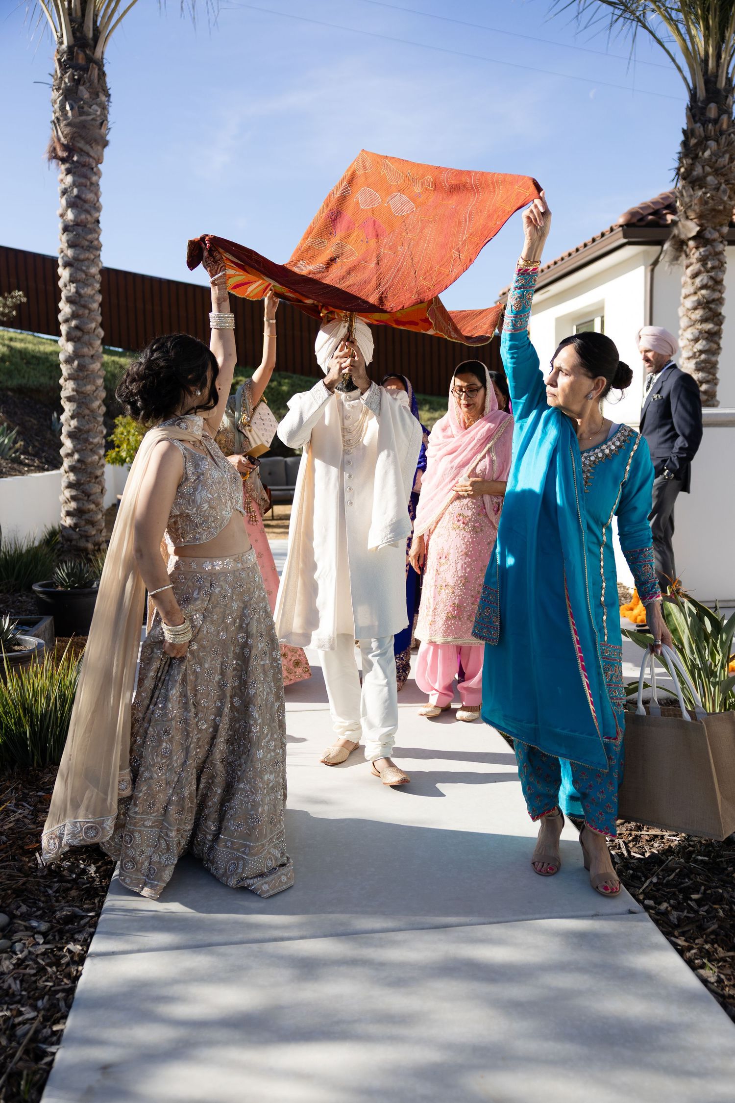 Traditional Indian wedding ceremony with guests gathered under an orange canopy between palm trees.
