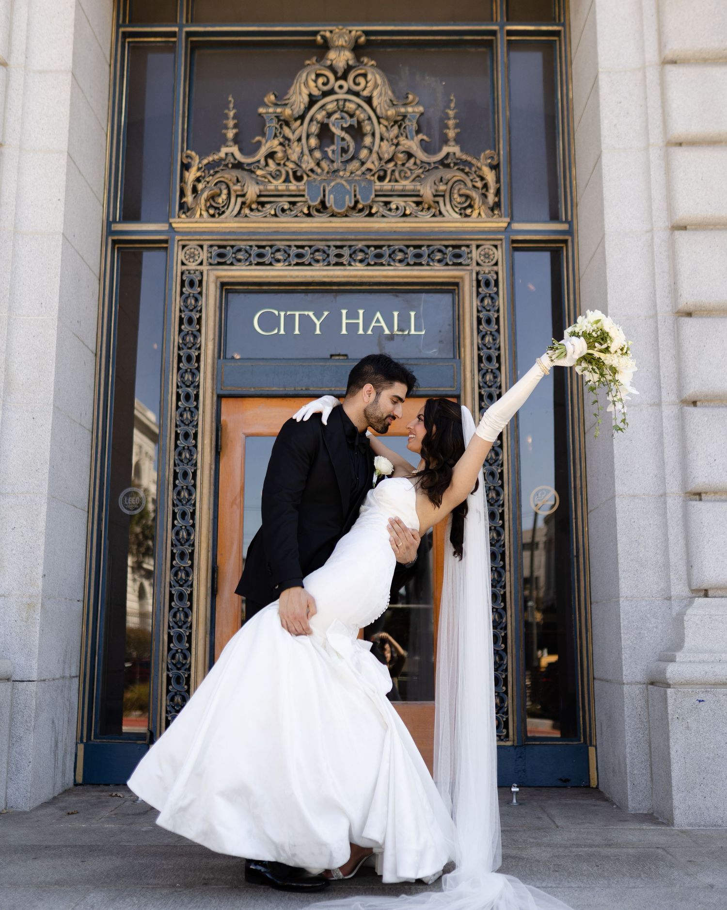 A romantic wedding couple shares a dip kiss in front of ornate City Hall doors with elegant architectural details.