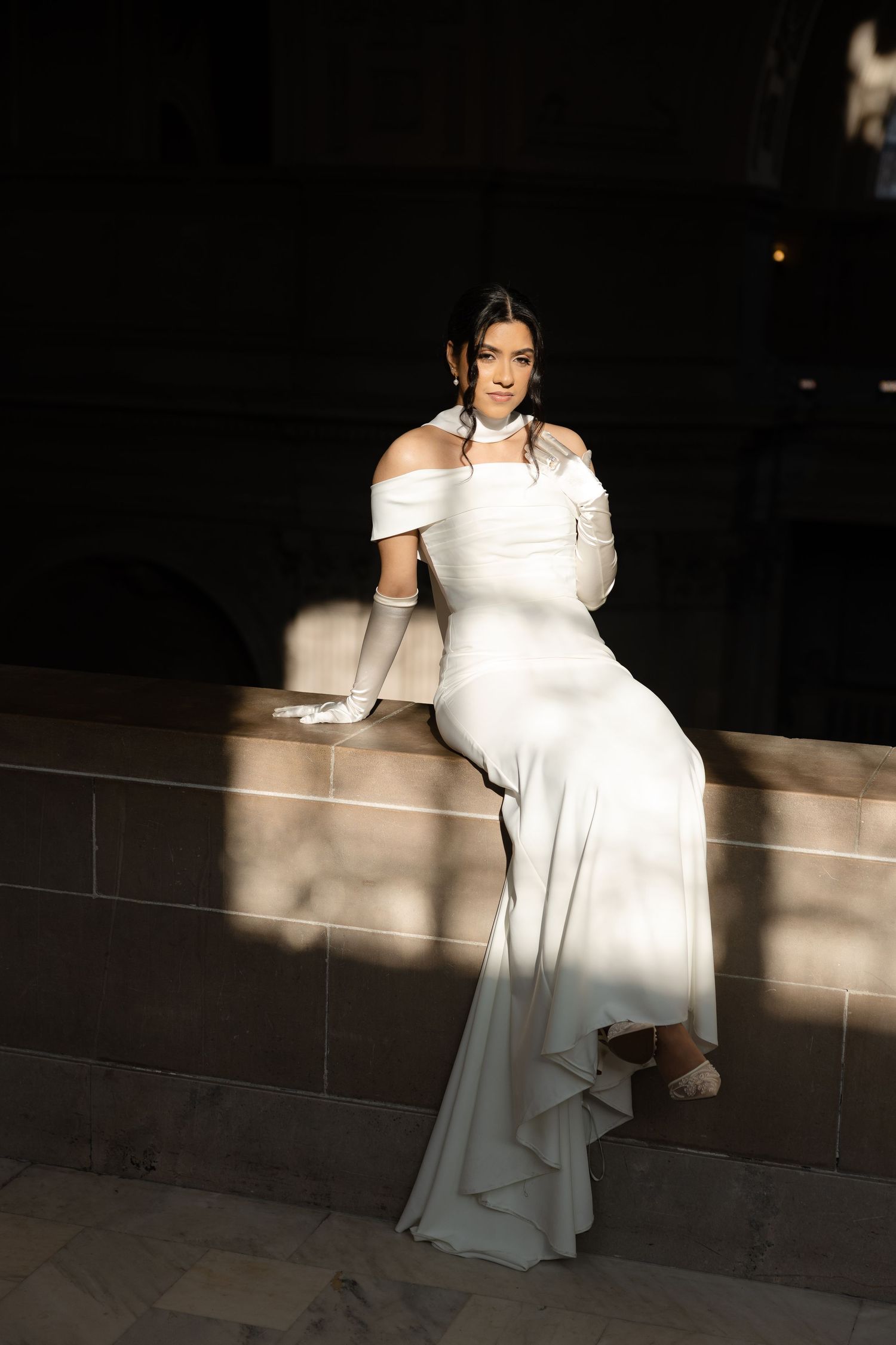 Elegant bride in off-shoulder white wedding gown and long gloves sits on stone ledge in dramatic lighting.