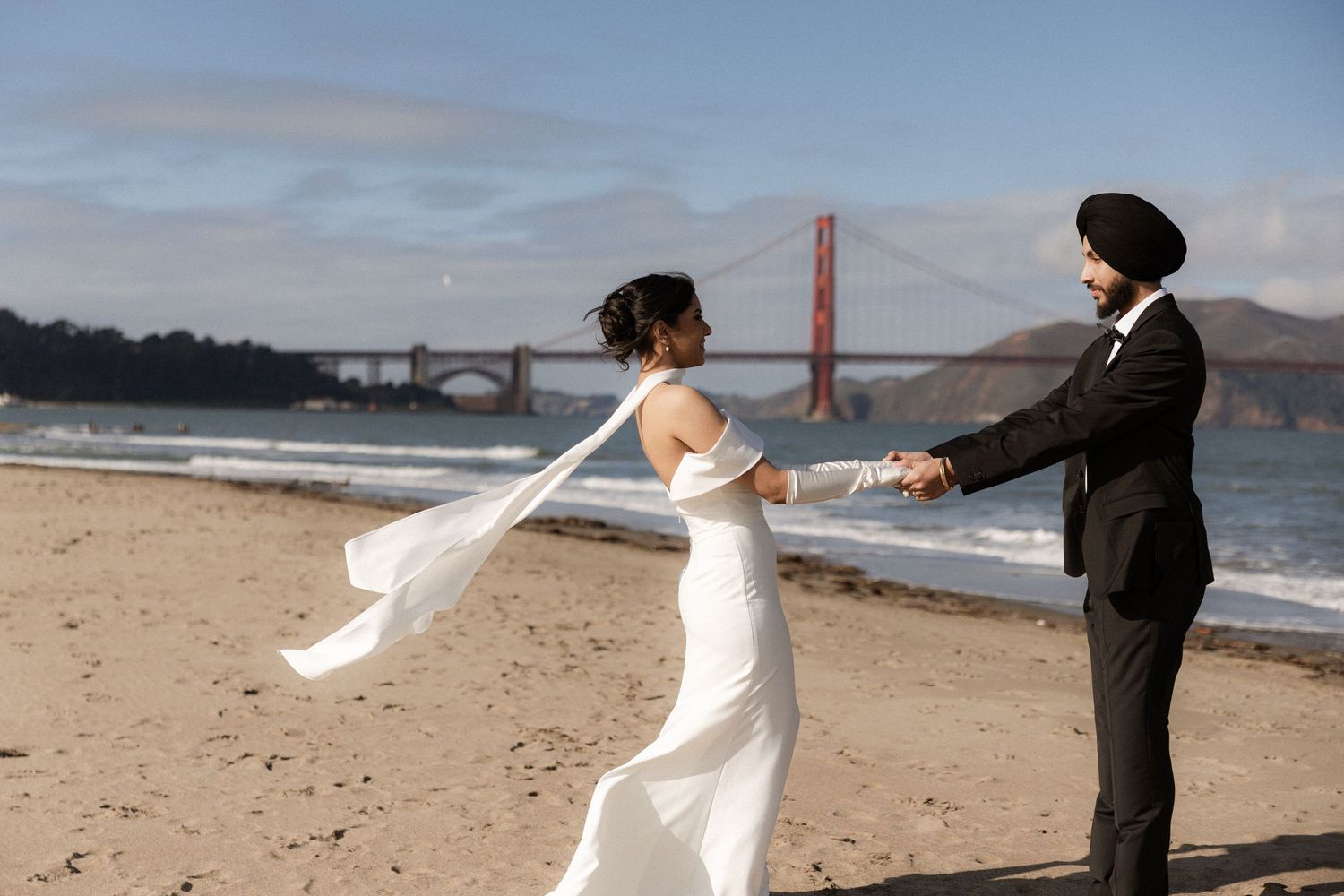A couple in wedding attire dances and spins joyfully on a sandy beach with the Golden Gate Bridge in the background.